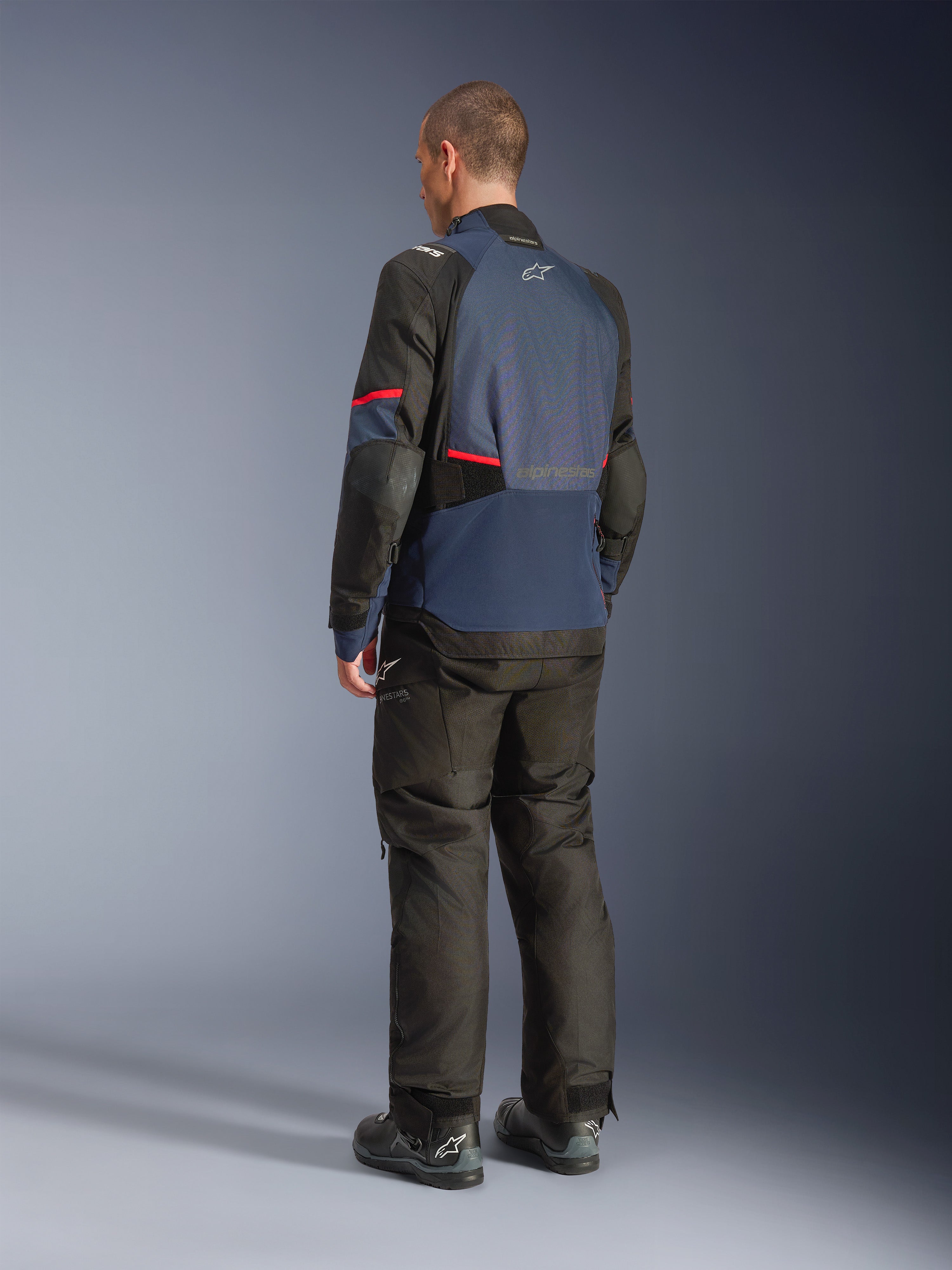 Alpinestars Andes V4 Drystar®-jacka, Night Navy och Mood Indigo med svarta paneler och röda accenter, vy snett bakifrån, bärs med matchande svarta äventyrsbyxor och stövlar, teknisk textilkonstruktion med Alpinestars-logotyp på övre delen av ryggen, förlängd fåll baktill för täckning.