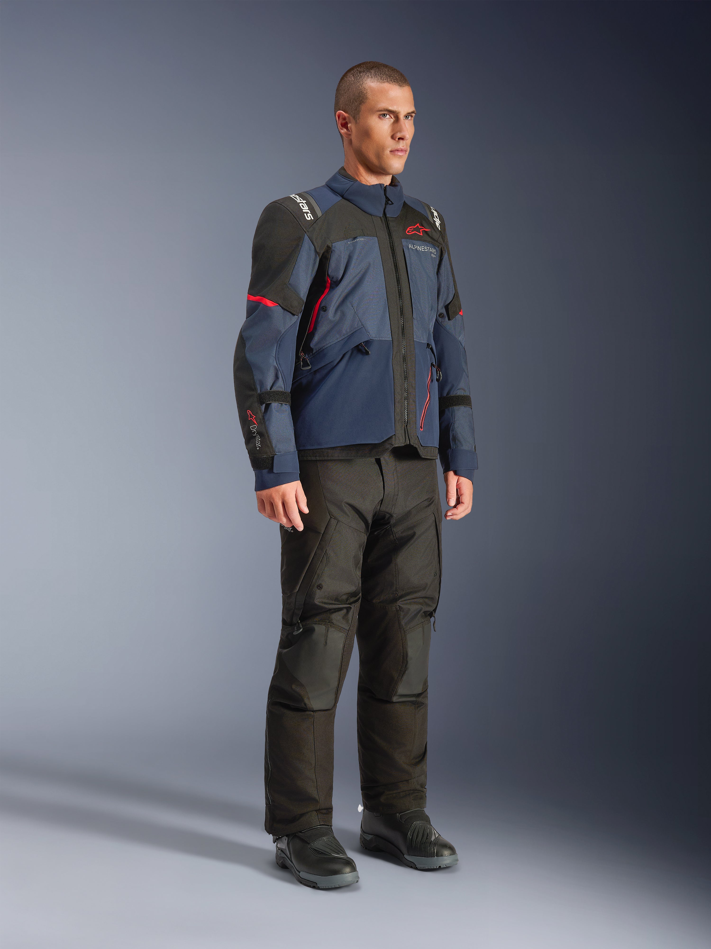 Manlig modell som bär Alpinestars Andes V4 Drystar®-jacka i Night Navy och Mood Indigo med svarta paneler och röda detaljer, kombinerat med svarta motorcykelbyxor och stövlar, teknisk adventure touring-utrustning i studiomiljö.