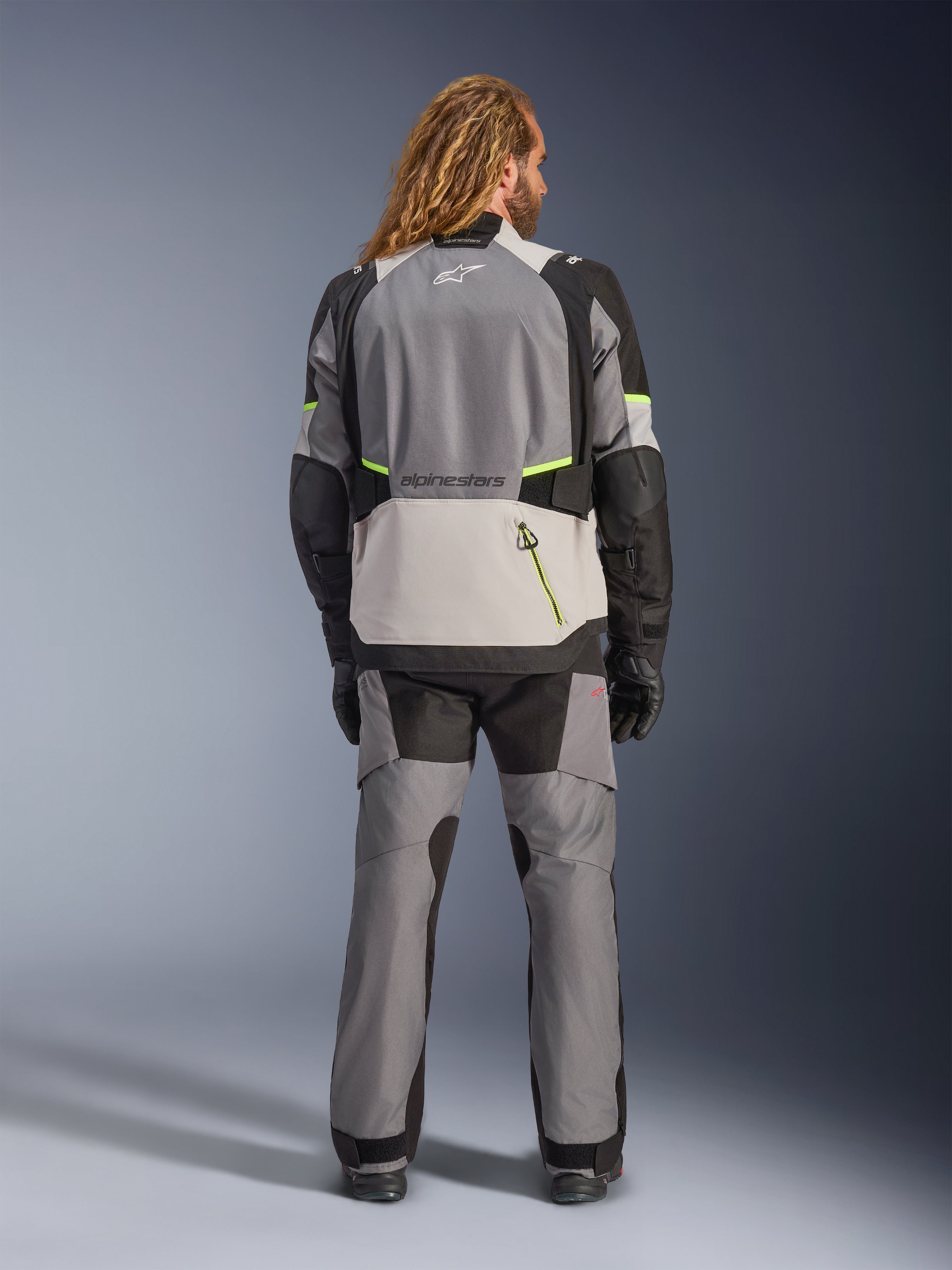 Alpinestars Andes V4 Drystar® jacka, äventyrsmotorcykeljacka, mörkgrå och isgrå med svarta och neongula accenter, vy bakifrån bärs med matchande byxor och svarta handskar, med en stor bakficka med gul dragkedja och förstärkta axel- och armbågspaneler