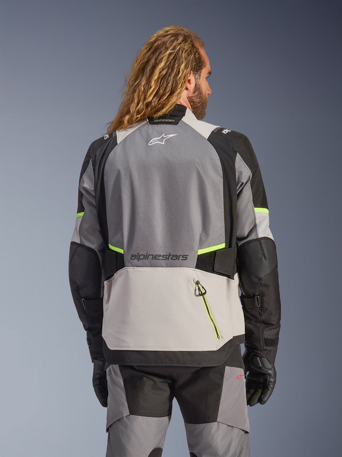 Alpinestars Andes V4 Drystar® jacka, vy bakifrån av äventyrsmotorcykeljacka i mörkgrått, isgrått och svart med fluorescerande gula accenter, med förstärkta textilpaneler och Alpinestars-branding, bärs med matchande mc-byxor och svarta handskar.