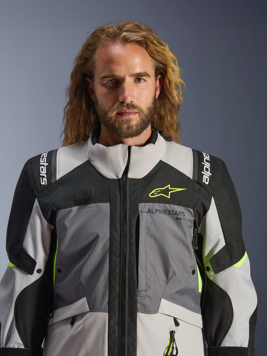 Alpinestars Andes V4 Drystar®-jacka, mörkgrå, isgrå, svart och neongul, bärs av en man med långt vågigt hår, teknisk konstruktion i flera material, Alpinestars-logotyp på bröstet och axlarna, hög krage för äventyrskörning