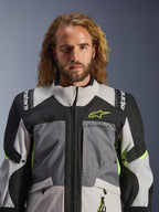 Alpinestars Andes V4 Drystar®-jacka, mörkgrå, isgrå, svart och neongul, bärs av en man med långt vågigt hår, teknisk konstruktion i flera material, Alpinestars-logotyp på bröstet och axlarna, hög krage för äventyrskörning