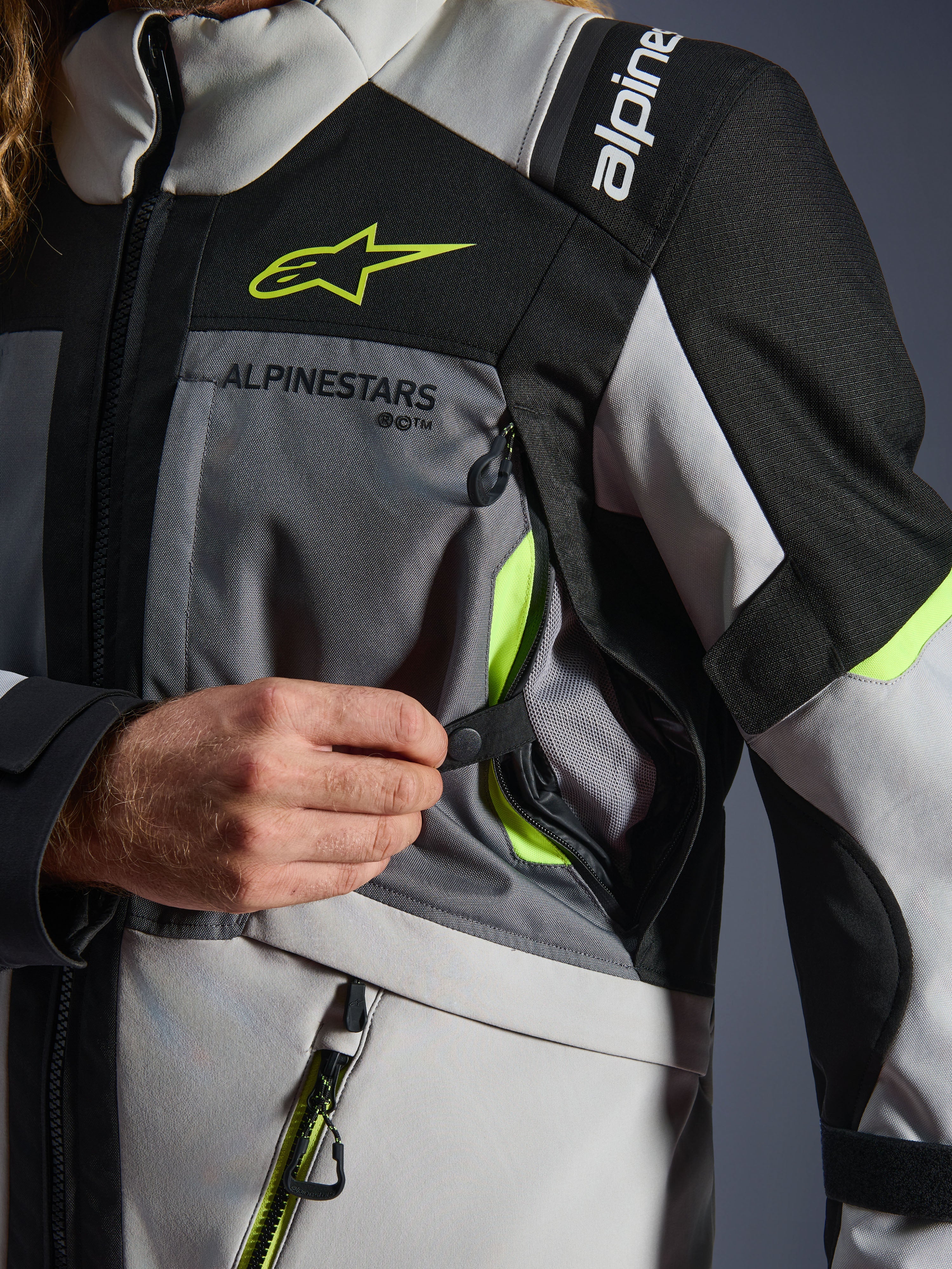 Alpinestars Andes V4 Drystar® jacka, motorcykeljacka, mörkgrå/isgrå/svart/fluorgul, närbild som visar en hand som öppnar ventilationsintaget på bröstet med neongula detaljer och nätfoder, slitstark textilkonstruktion med Alpinestars-branding.