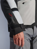 Alpinestars Andes V4 Drystar® jacka, äventyrsmotorcykeljacka, paneler i Ice Gray och svart, närbild på ärm med röd Drystar®-logotyp och justerbara remmar, funktionell textildesign för körning i alla väder