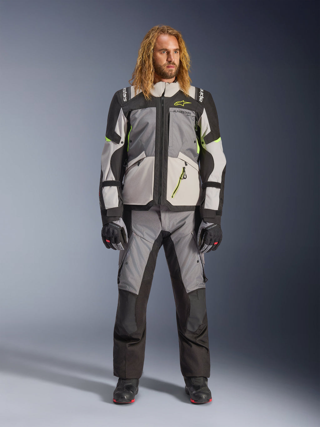 Manlig modell med långt blont hår i Andes V4 Drystar®-jacka i mörkgrått, isgrått, svart och gul fluo, kombinerat med matchande äventyrsmotorcykelbyxor och svarta stövlar, komplett teknisk körutrustning för touring i alla väder.