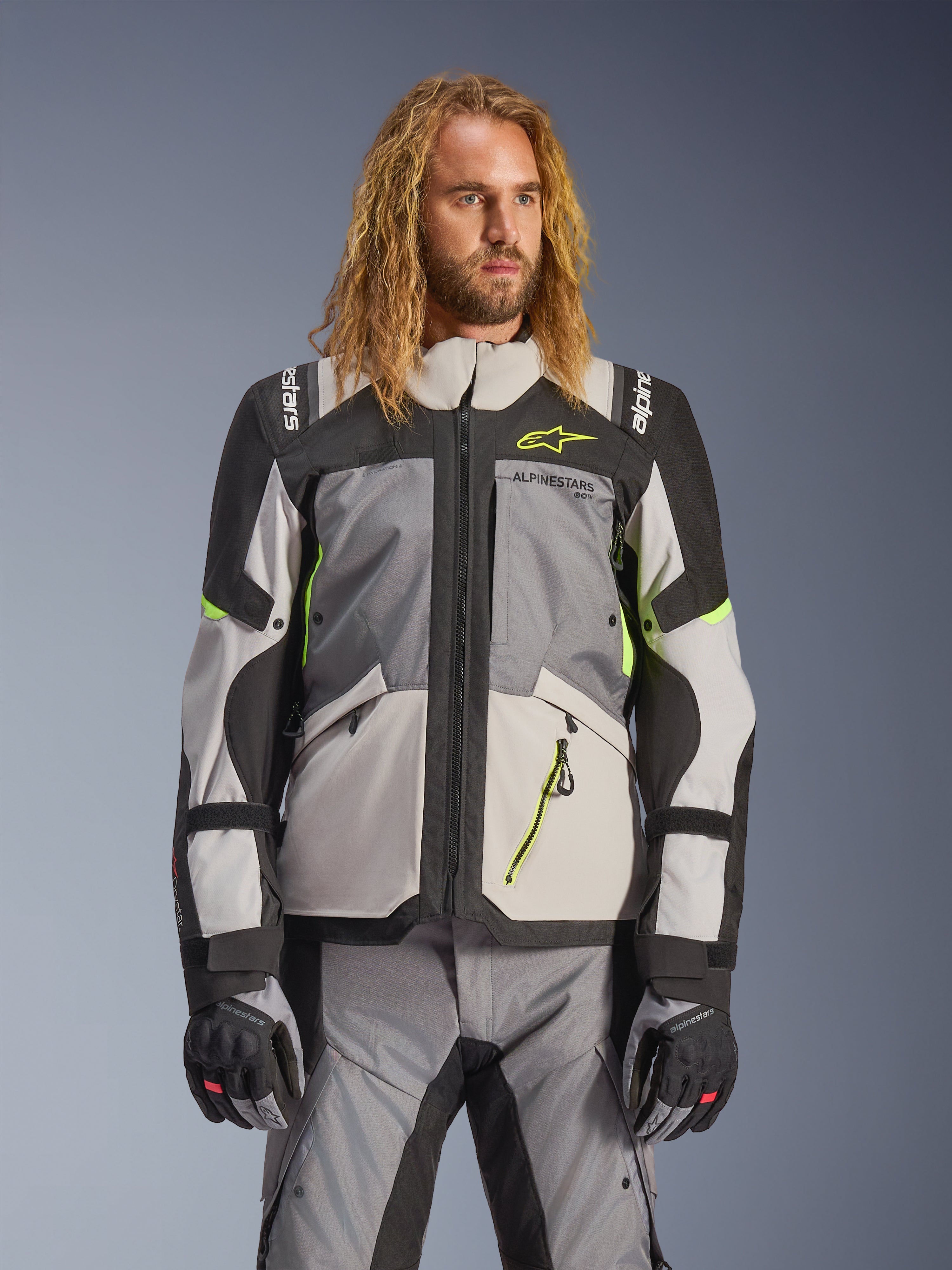Alpinestars Andes V4 Drystar-jacka, äventyrs- och touringjacka för motorcykel, mörkgrå och isgrå med neongula detaljer, bärs av en modell med matchande byxor och handskar, textilkonstruktion med flera fickor för körning i alla väder