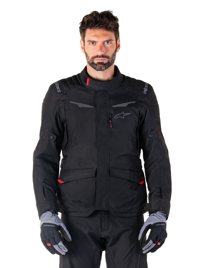 Alpinestars ST-1 Waterproof-jacka, svart adventure touring-jacka för motorcykel, bärs av en manlig modell med matchande svarta byxor och grå/svarta handskar, minimalistisk styling, Alpinestars-logotyp på axlarna, stora främre cargo-fickor med röda detaljer, ergonomisk konstruktion för vägtouring i alla väder.