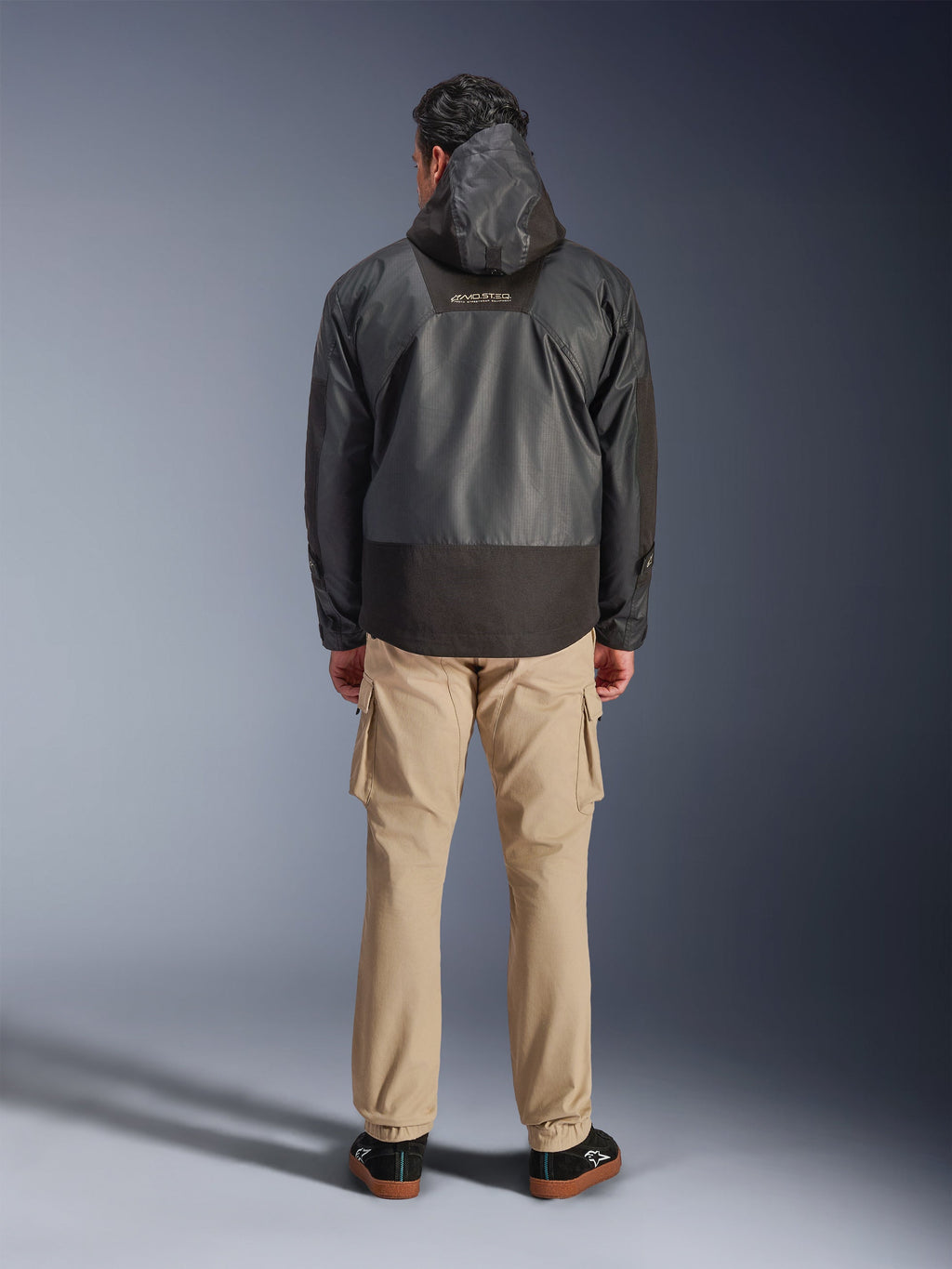 Alpinestars Mospher Waterproof Jacket, svart motorcykelparkas sedd bakifrån, med MO.ST.EQ-logotyp på övre delen av ryggen och ripstop-tyg, bärs med beige cargobyxor och svarta sneakers, urban pendlarstil.