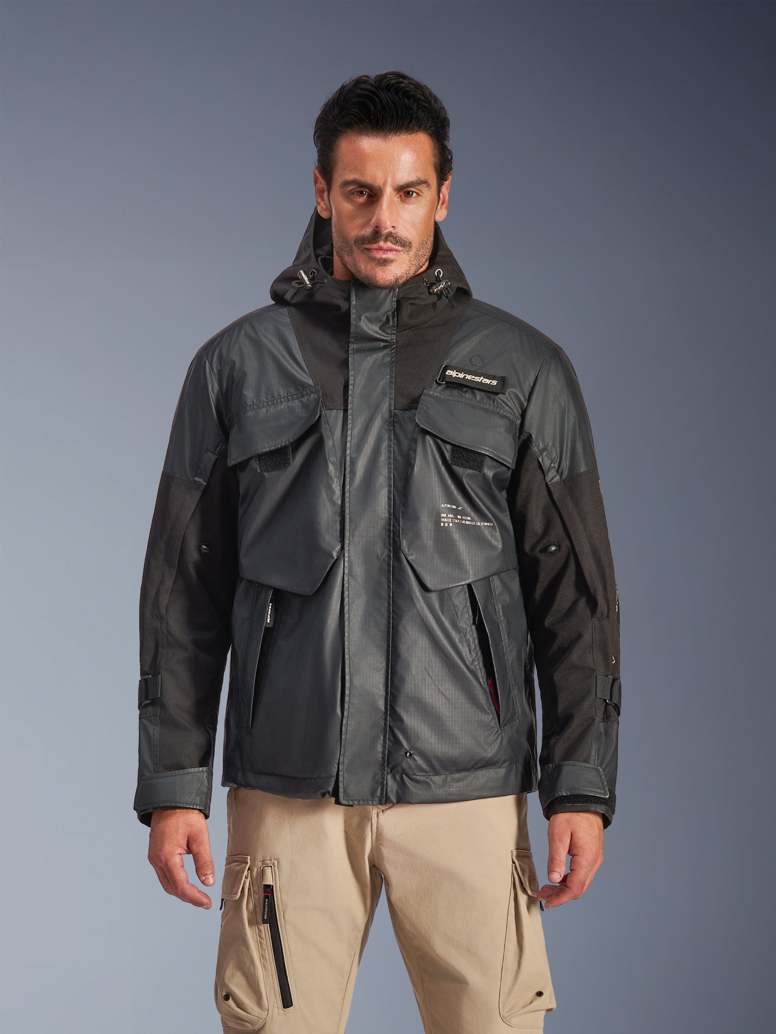 Alpinestars Mospher Waterproof Jacket, urban motorcykelparkas för män, svart och mörkgrått ripstop-tyg, design med huva och flera stora funktionella fickor, bärs av en modell med khakifärgade cargobyxor för en taktisk look