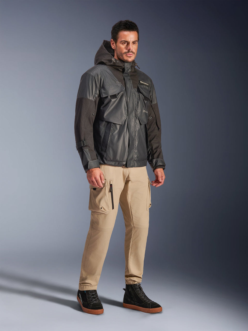 Manlig modell bär Alpinestars Mospher Waterproof Jacket i svart, en utility-inspirerad vattentät parka med huva och flera fickor, matchad med beige cargobyxor och svarta high-top sneakers, urban motorcykelutrustning för pendlare.