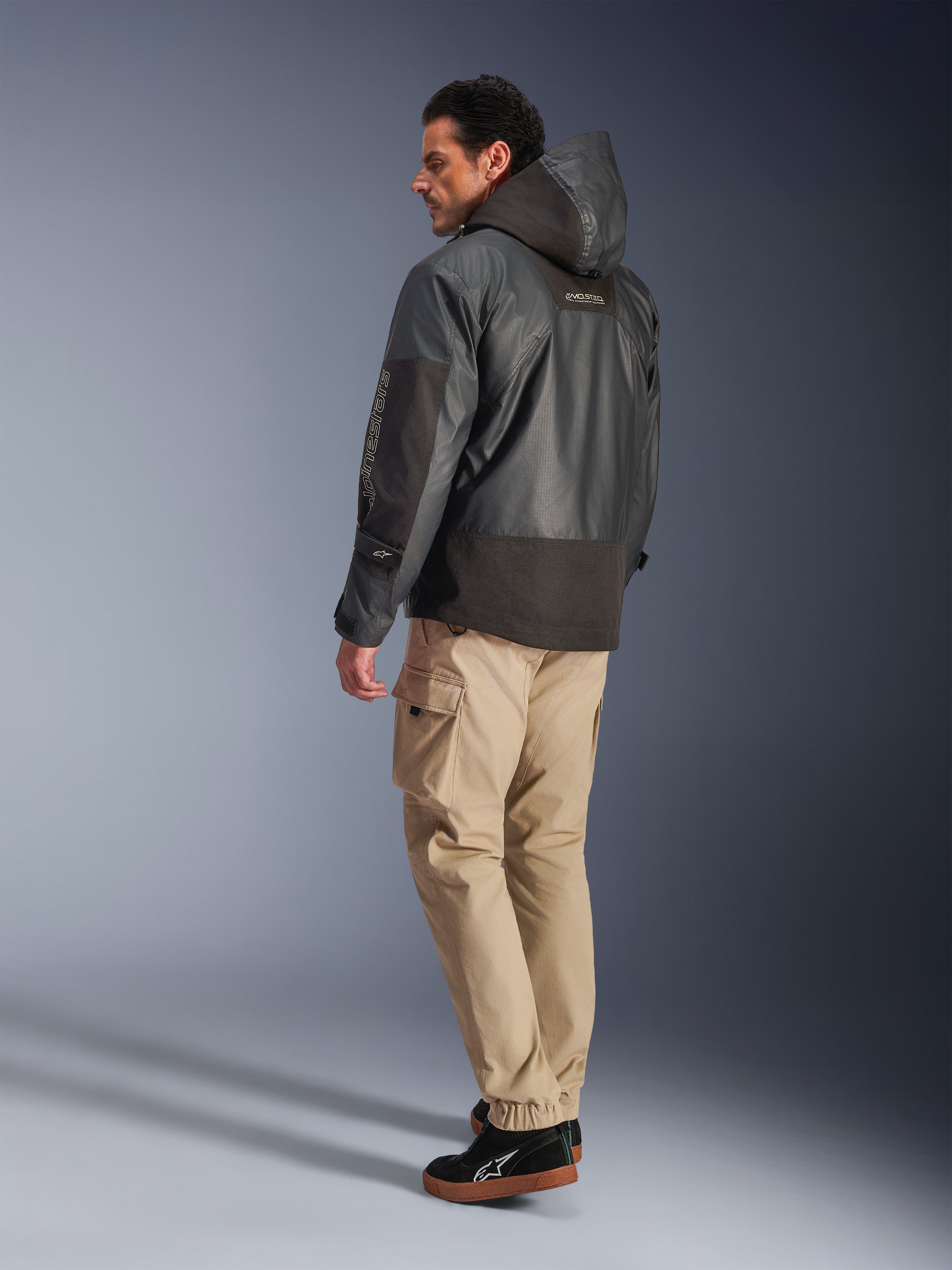 Man som bär Alpinestars Mospher Waterproof Jacket i svart, vy bakifrån och från sidan, kombinerad med beiga cargobyxor och svarta sneakers. Har en parkasdesign med huva i ripstop-tyg, Alpinestars-logotyp på ärmen och en modern urban motorcykelstil.
