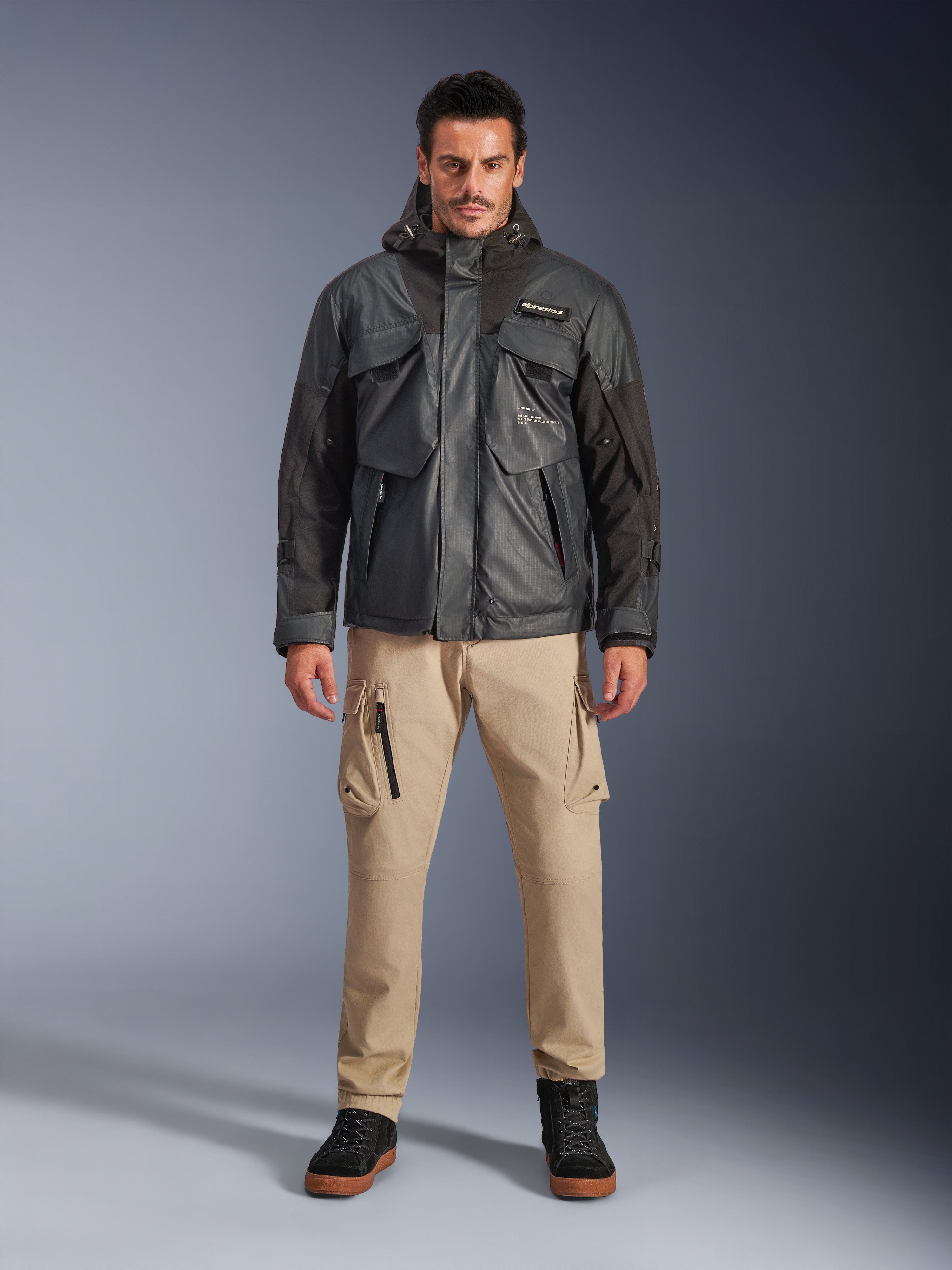 Manlig modell i Alpinestars Mospher Waterproof Jacket i svart, urban modern motorcykelutrustning, taktisk utility-design med flera funktionella fickor och huva, kombinerat med beige cargobyxor och svarta skor, helkroppsporträtt i studio.