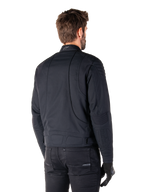 Alpinestars Clayton WR Jacket in Black, rear three-quarter view on a male model, urban motorcycle jacket with quilted padding on shoulders and elbows, technical stitch lines, and reflective logo on the collar, styled with black pants and gloves.
