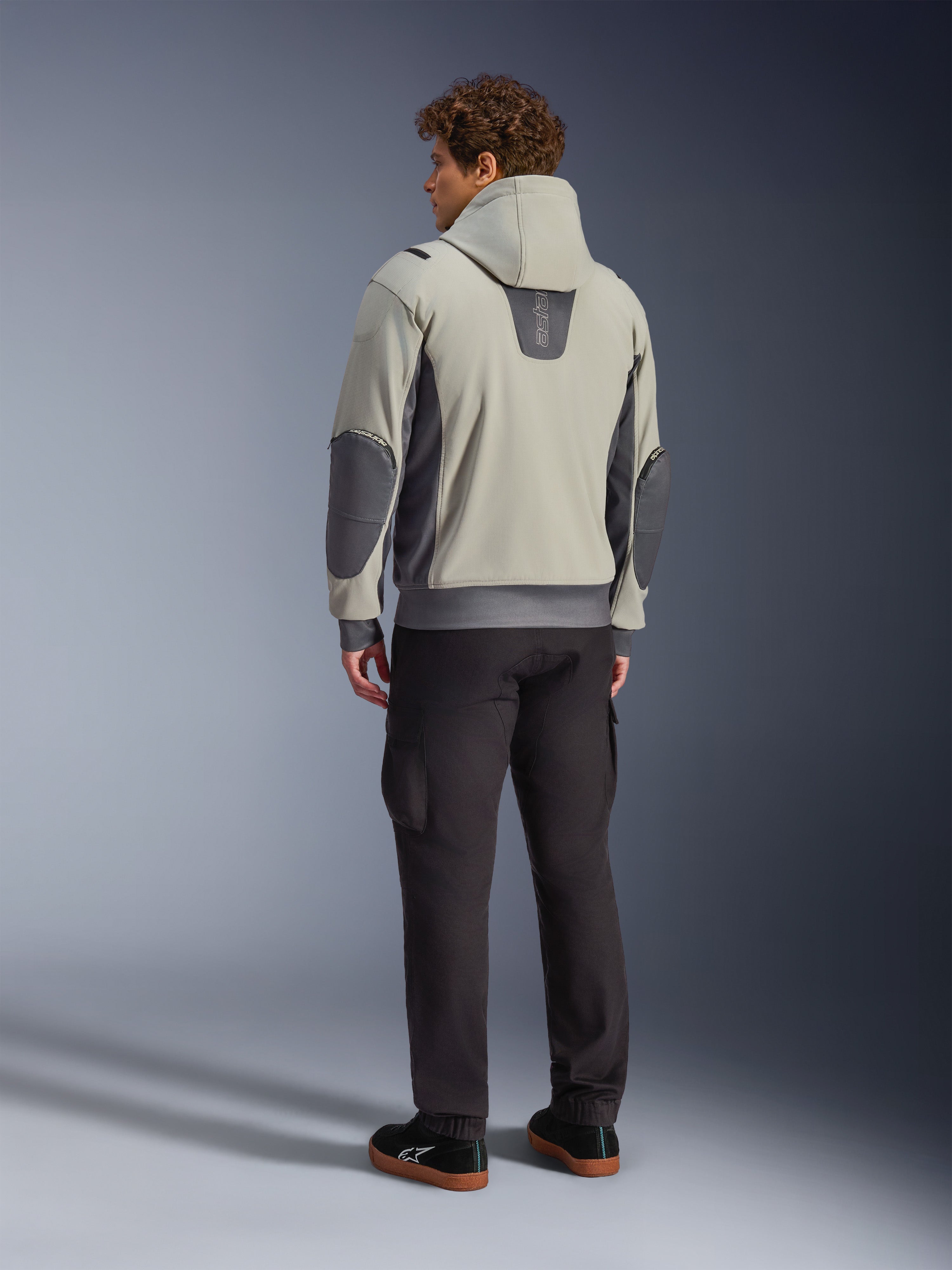 Male model viewed from the back wearing the Alpinestars Moblast Waterproof Jacket in Iron Gray Dark Sage, hooded urban motorcycle jacket with dark gray side panels and reinforced elbow patches, paired with dark riding pants and black sneakers, modern minimalist aesthetic for urban commuting.