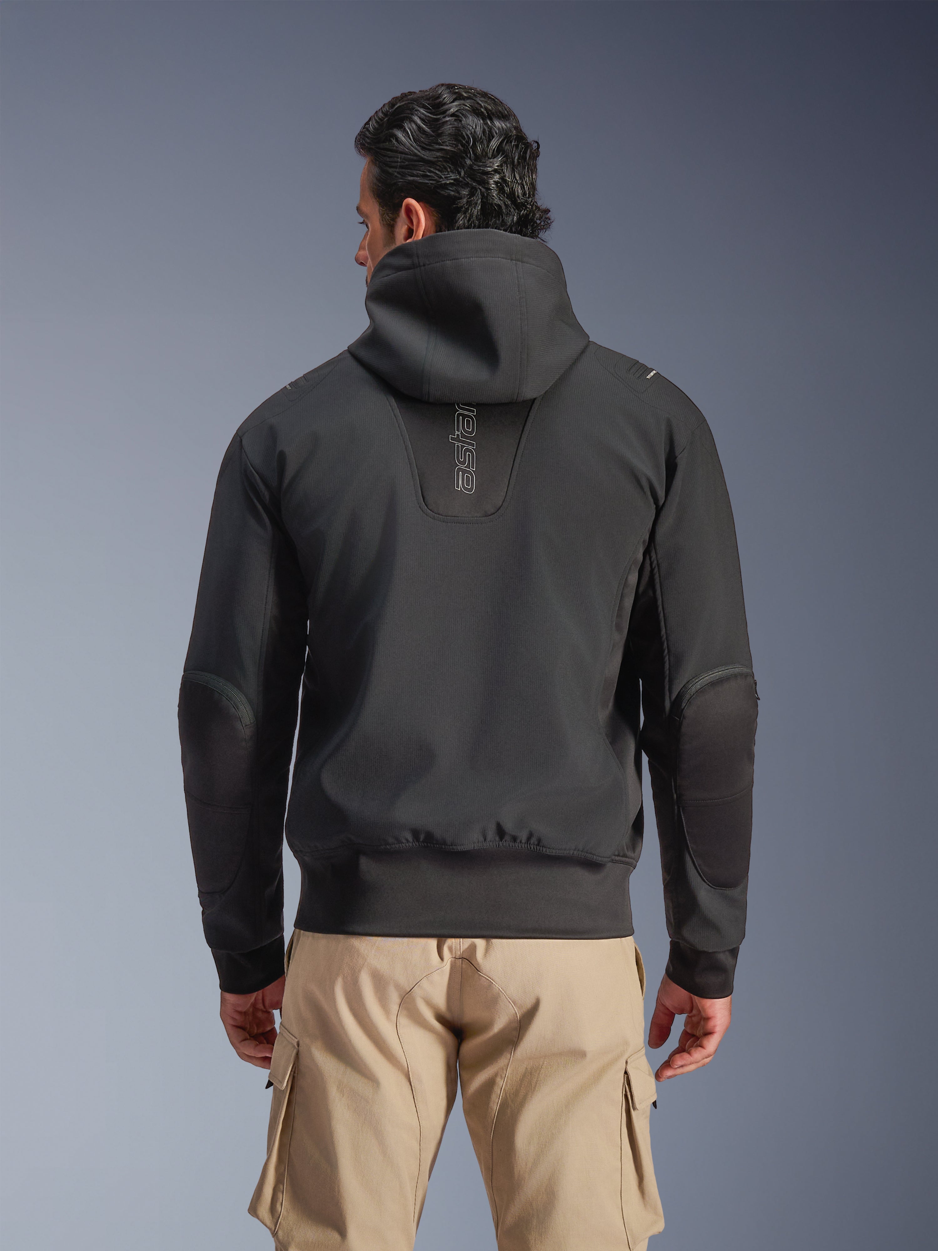 Alpinestars Moblast Waterproof Jacket, Black, Rear view of urban hooded motorcycle jacket, featuring vertical 'astars' logo on the back, elasticated waist and cuffs, reinforced elbows, and modern tech-forward design, worn with tan cargo pants.