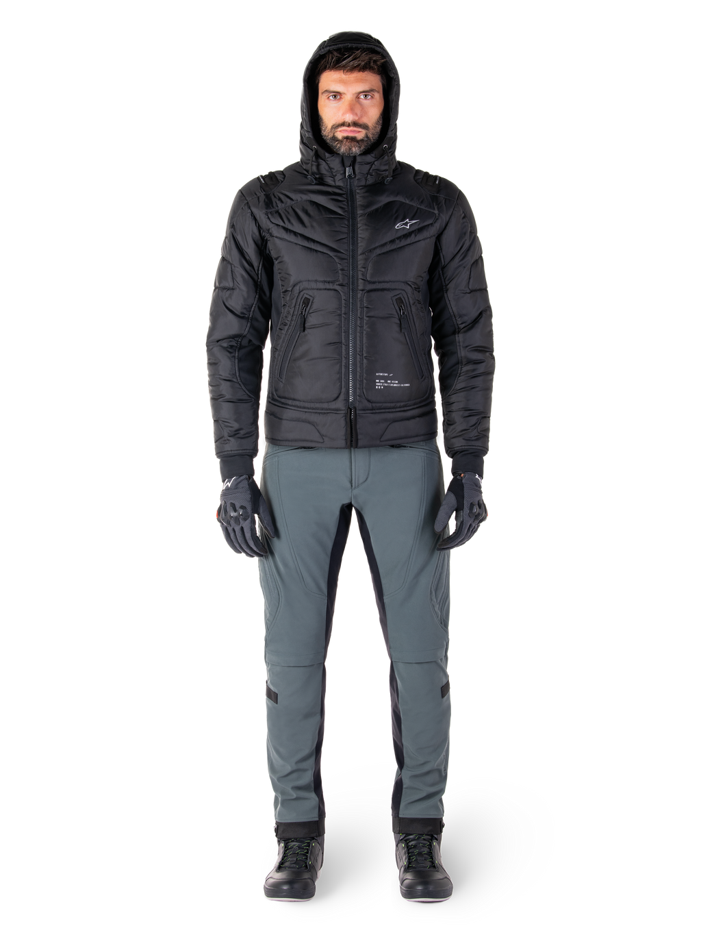 Alpinestars Mohobbs Waterproof Jacket i svart, manlig modell som bär en quiltad vadderad bomberjacka med huva, matchad med grå MC-byxor, svarta handskar och svarta stövlar, urban modern motorcykelutrustning.