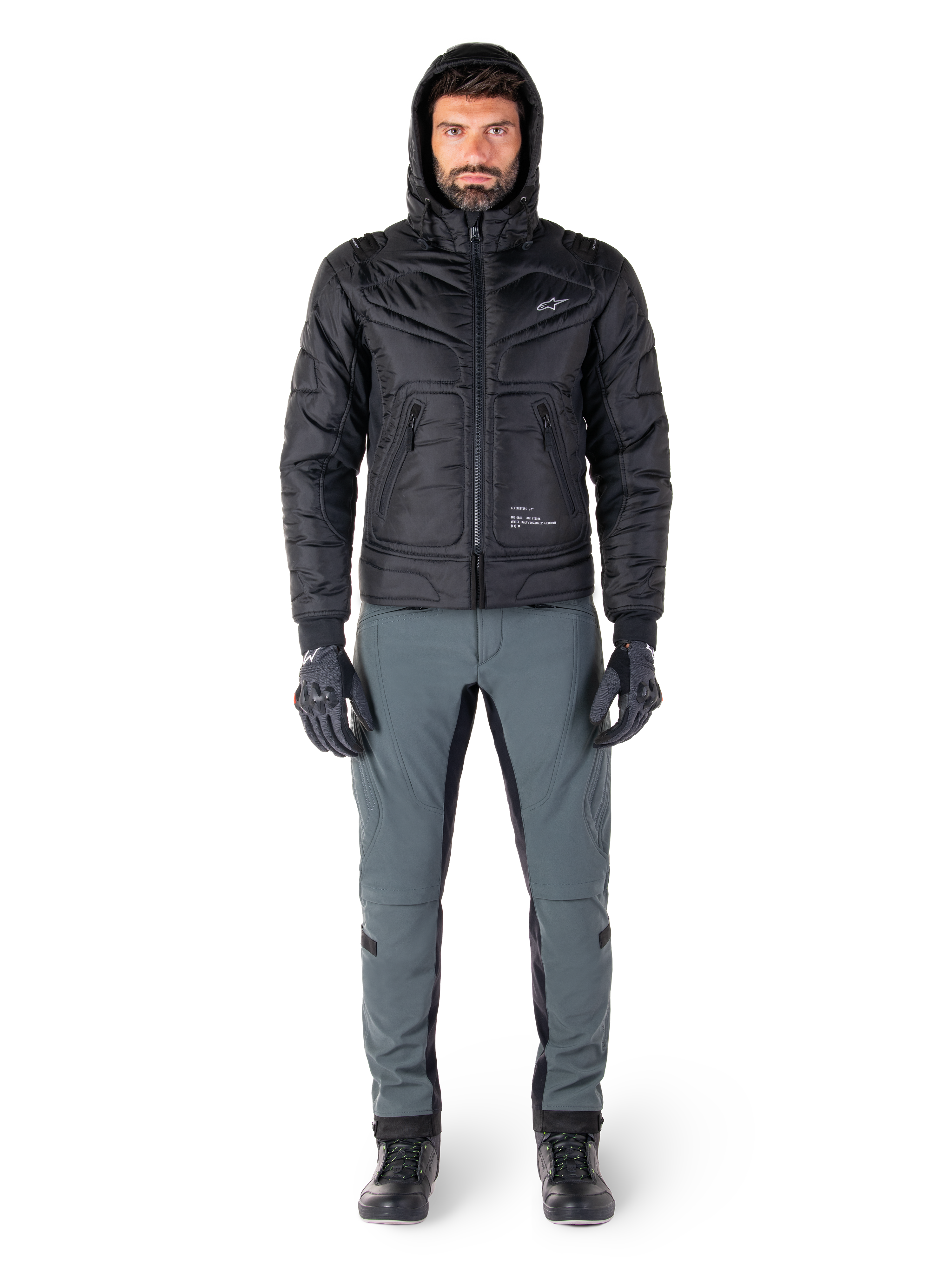 Alpinestars Mohobbs Waterproof Jacket i svart, manlig modell som bär en quiltad vadderad bomberjacka med huva, matchad med grå MC-byxor, svarta handskar och svarta stövlar, urban modern motorcykelutrustning.