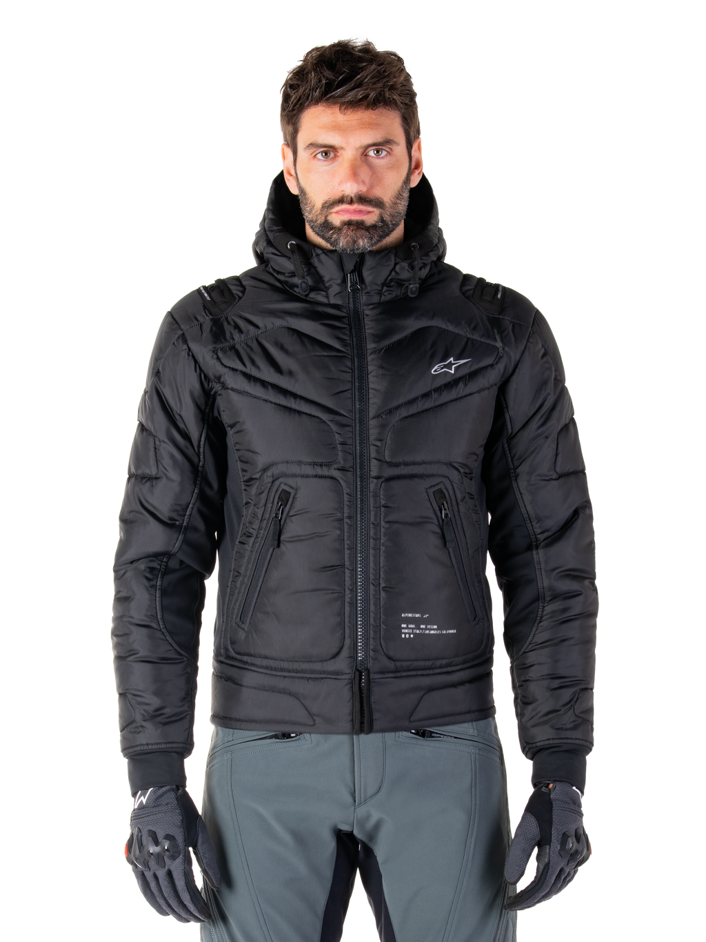 Alpinestars Mohobbs Waterproof Jacket, svart vadderad bomberjacka för motorcykel, quiltat polytyg med dragkedja fram och fickor med dragkedja, liten vit logotyp på bröstet, bärs av en modell med gråa byxor och handskar, urban streetwear-design för stadskörning.