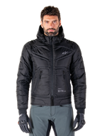 Alpinestars Mohobbs Waterproof Jacket, svart vadderad bomberjacka för motorcykel, quiltat polytyg med dragkedja fram och fickor med dragkedja, liten vit logotyp på bröstet, bärs av en modell med gråa byxor och handskar, urban streetwear-design för stadskörning.
