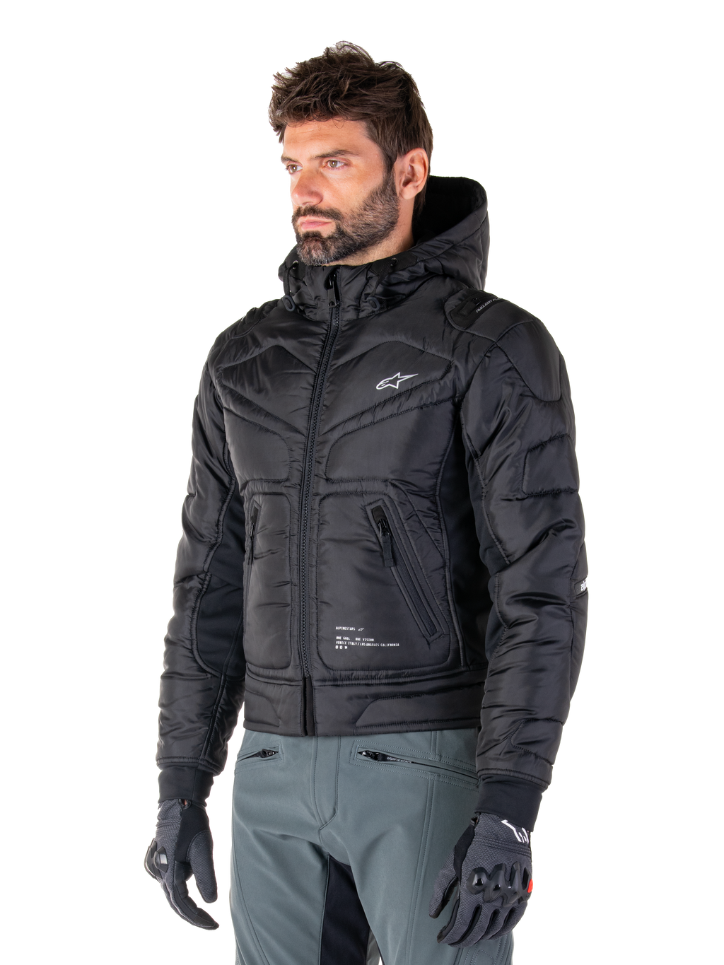 Alpinestars Mohobbs Waterproof Jacket, svart, quiltad vadderad bomberdesign med huva, bärs av en man med grå körbyxor och svarta handskar, urban streetwear-inspirerade motorcykelkläder med en liten vit logotyp på bröstet.