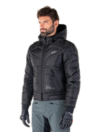 Alpinestars Mohobbs Waterproof Jacket, svart, quiltad vadderad bomberdesign med huva, bärs av en man med grå körbyxor och svarta handskar, urban streetwear-inspirerade motorcykelkläder med en liten vit logotyp på bröstet.