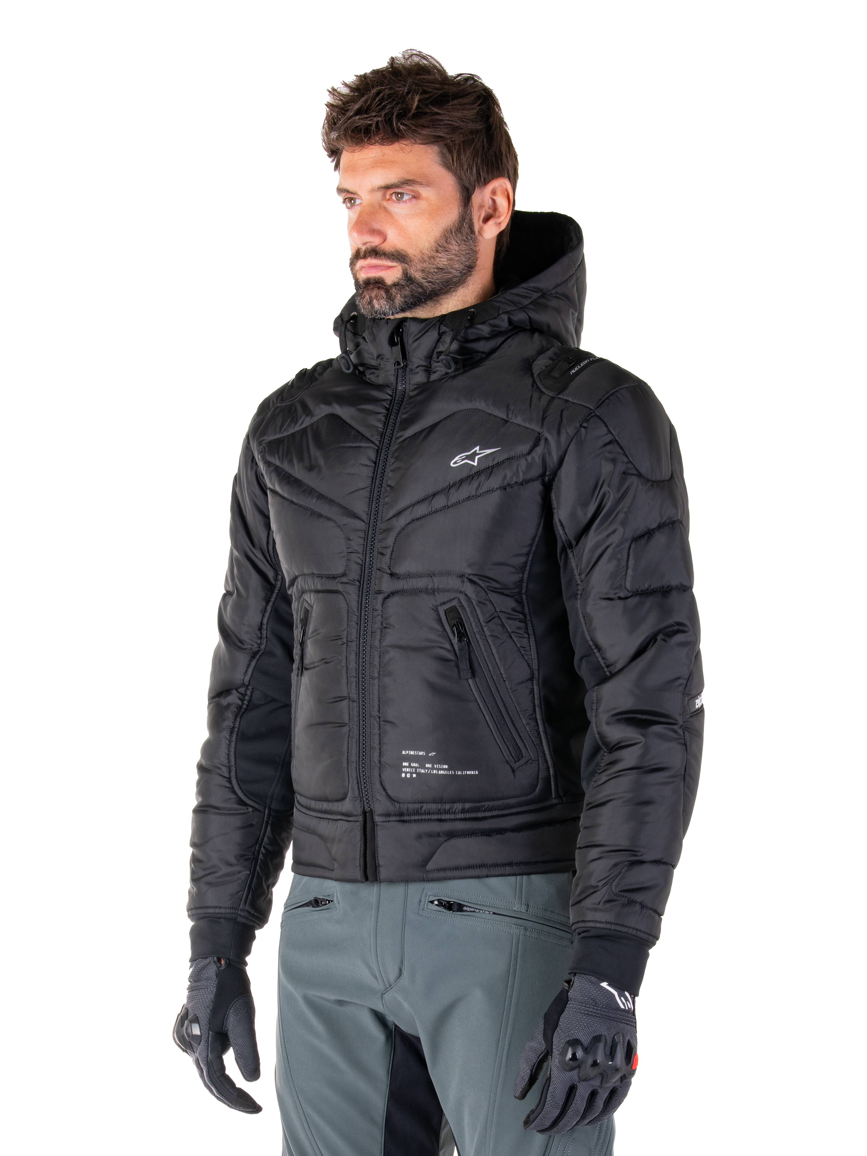 Alpinestars Mohobbs Waterproof Jacket, svart, quiltad vadderad bomberdesign med huva, bärs av en man med grå körbyxor och svarta handskar, urban streetwear-inspirerade motorcykelkläder med en liten vit logotyp på bröstet.