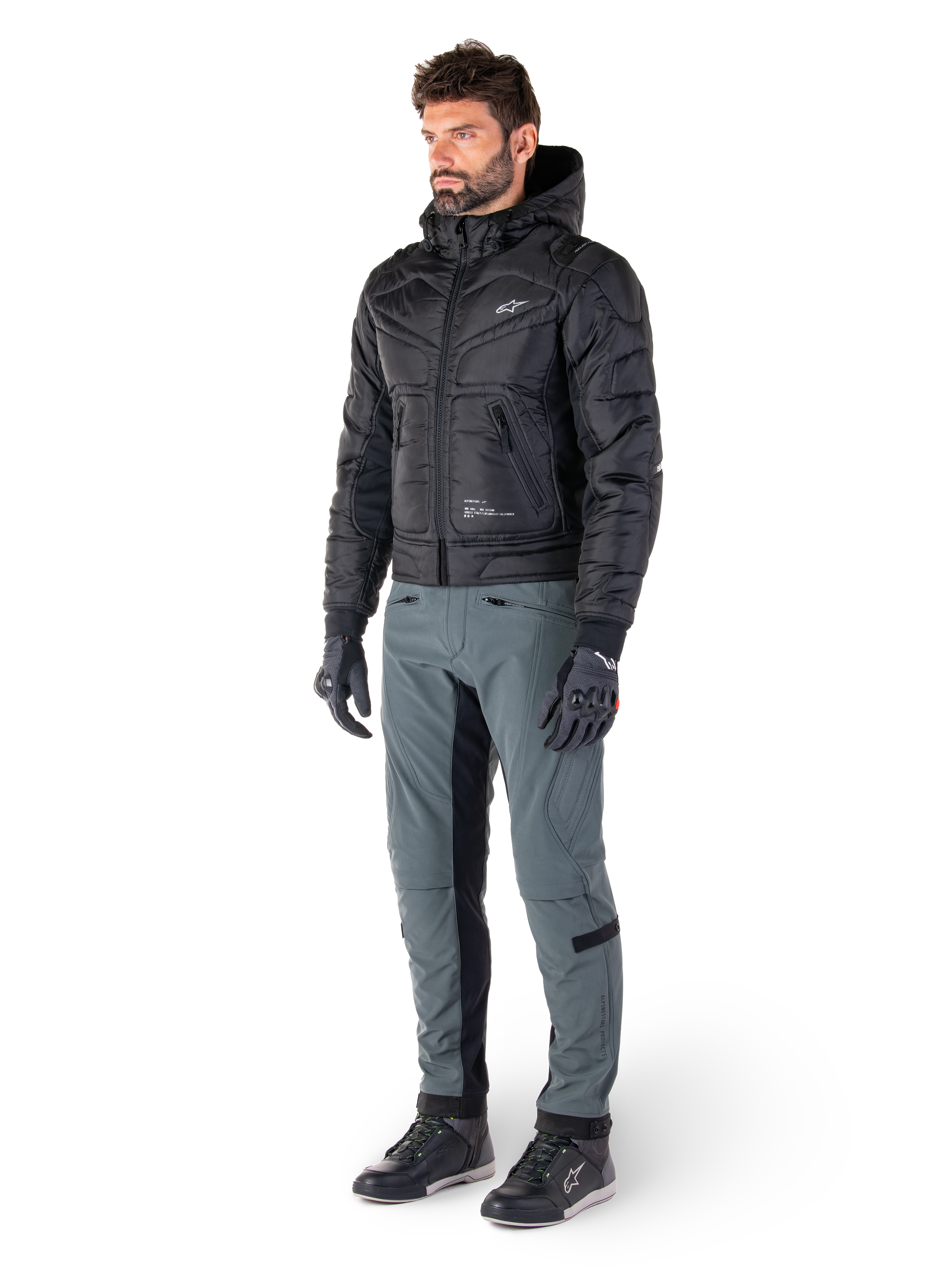 Alpinestars Mohobbs Waterproof Jacket, svart, quiltad urban bomberjacka för motorcykel med huva, bärs av en modell med grå körbyxor och svarta handskar, med vit logotyp på bröstet, modern streetwear-inspirerad skyddsutrustning