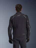 Alpinestars Andes Pro Drystar® XF-jacka, svart och mörkgrå, baksida buren av en manlig modell, med ett stort bakre förvaringsfack, reflexdetaljer på axlarna och logotypmärkning, kombinerad med svarta handskar och byxor för äventyrskörning.