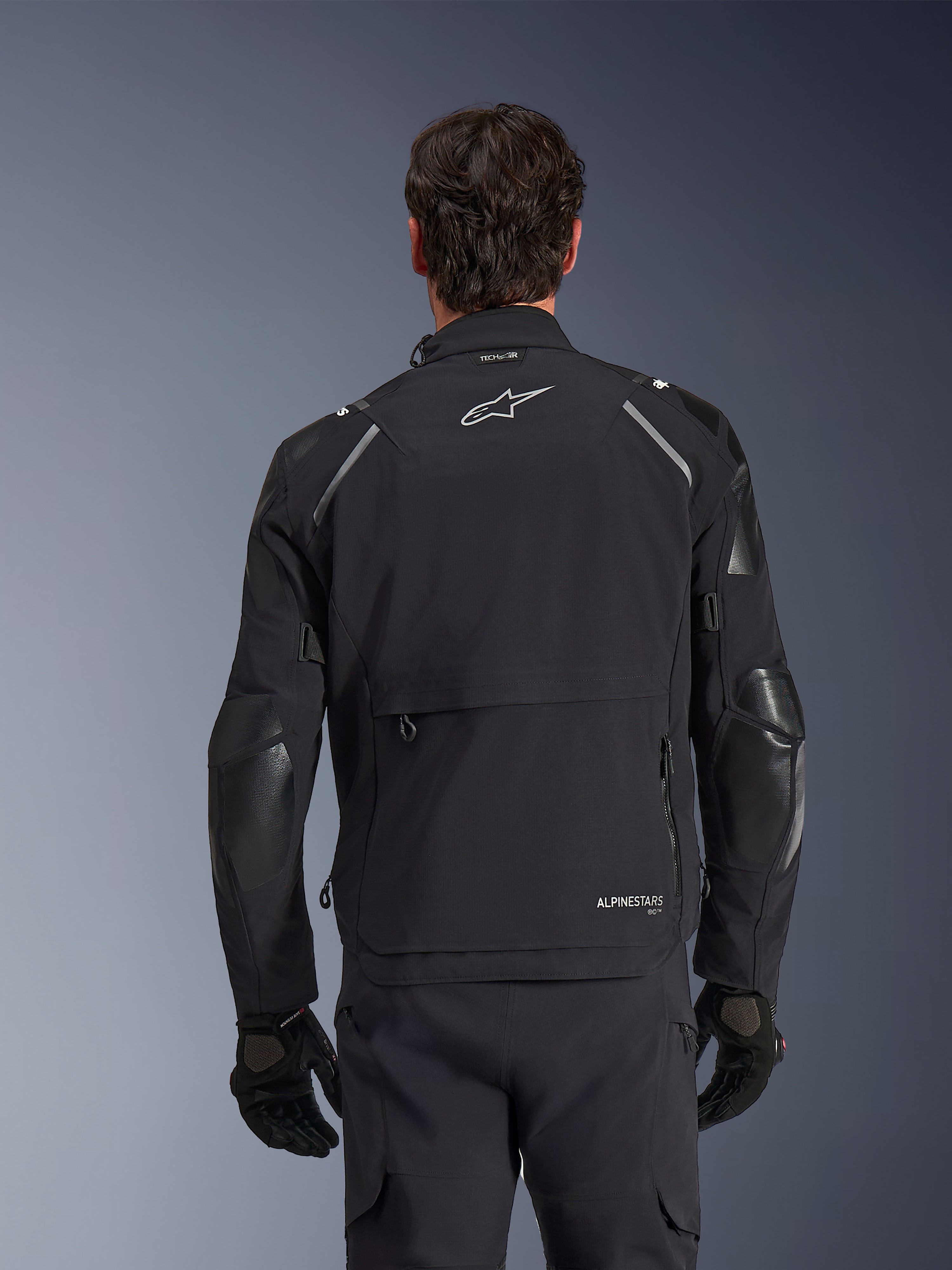 Alpinestars Andes Pro Drystar® XF-jacka, svart och mörkgrå, baksida buren av en manlig modell, med ett stort bakre förvaringsfack, reflexdetaljer på axlarna och logotypmärkning, kombinerad med svarta handskar och byxor för äventyrskörning.