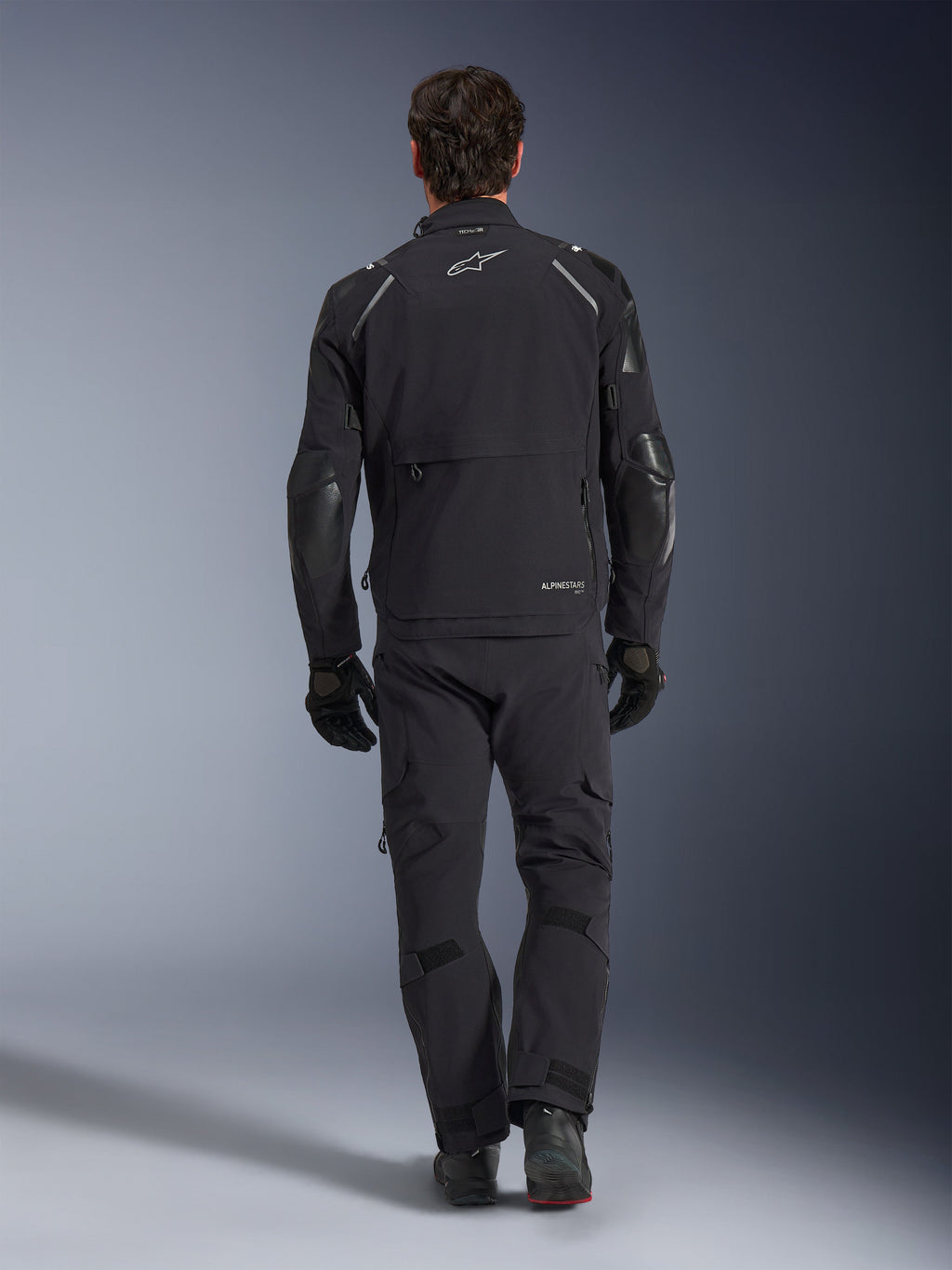 Alpinestars Andes Pro Drystar® XF-jacka, äventyrsjacka för touring, svart och mörkgrå, vy bakifrån som visar en stor cargoficka och förstärkta armbågspaneler, bärs med matchande svarta byxor och handskar, stilren funktionell design för körning i alla väder