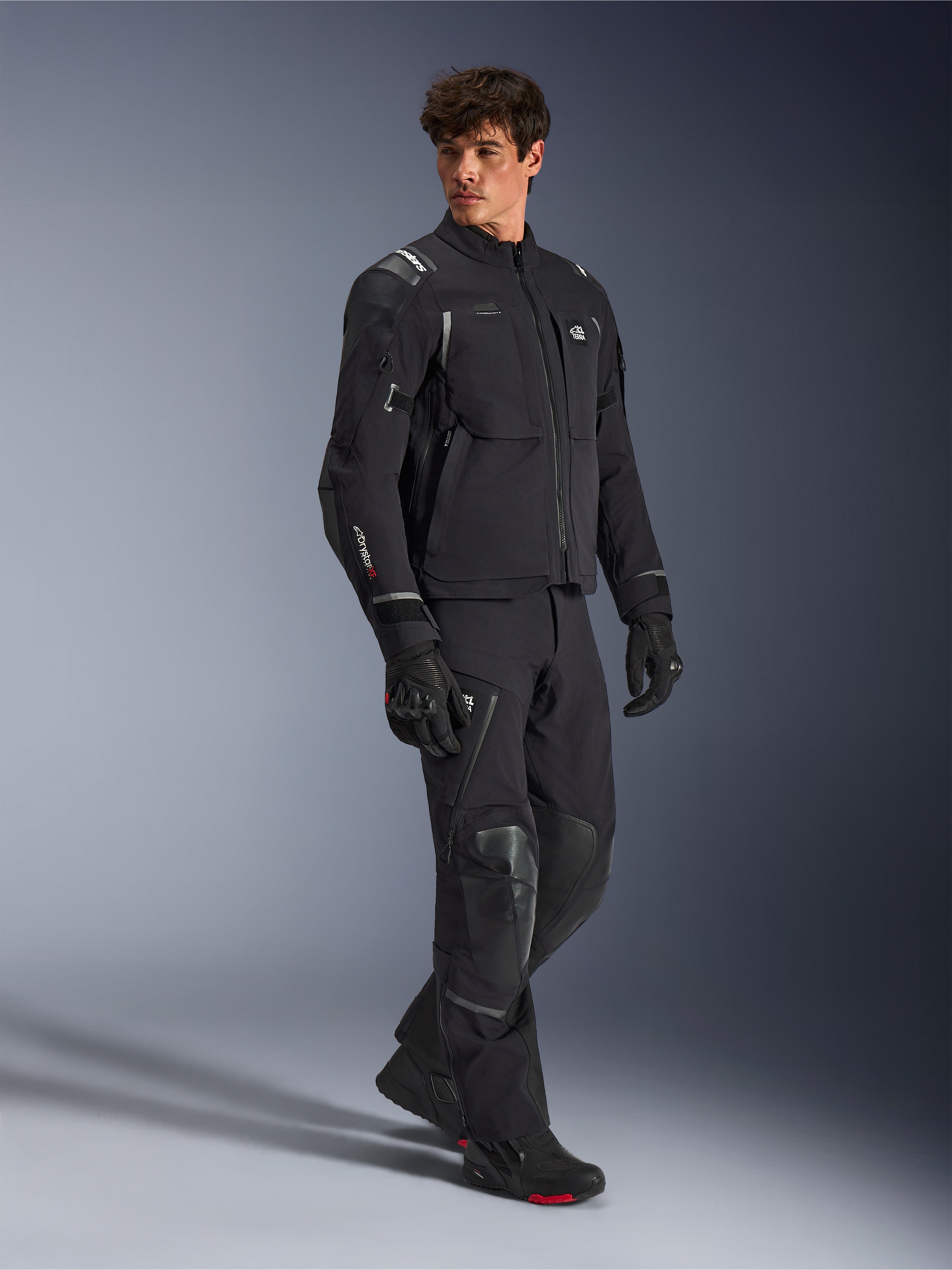 Alpinestars Andes Pro Drystar® XF-jacka, svart och mörkgrå motorcykeljacka, bärs av manlig modell med matchande äventyrsbyxor, handskar och stövlar, helkroppsbild i studio, teknisk äventyrsutrustning för touring med reflexdetaljer och logotyper