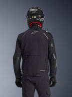 Alpinestars Andes Pro Drystar® XF Jacket i svart, ryggvy på en förare som bär en kolfiberhjälm, med ett stort bakre förvaringsfack och vit Alpinestars-logotyp, teknisk allvädersutrustning för äventyrsmotorcykelkörning.