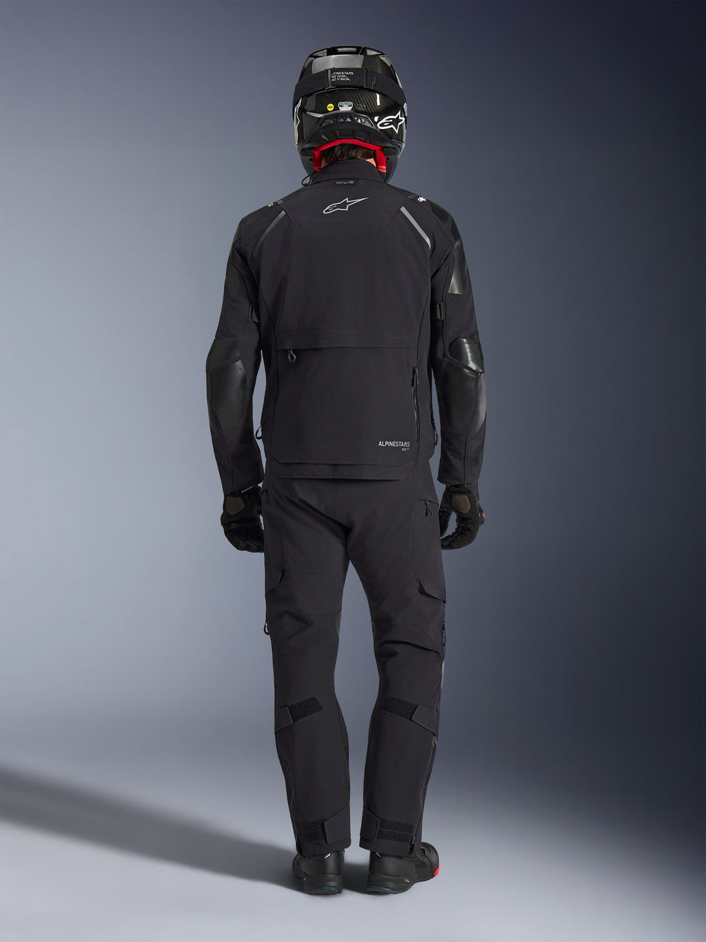 Alpinestars Andes Pro Drystar® XF-jacka i svart och mörkgrå, förare sedd bakifrån i komplett adventure touring-utrustning inklusive matchande byxor och kolfiberhjälm, med en stor bakre förvaringsficka och reflekterande Alpinestars-logotyp på ryggen.