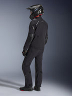 Motorcyklist klädd i Alpinestars Andes Pro Drystar® XF-jacka och matchande byxor i svart och mörkgrå, profilvy bakifrån, med en svart äventyrshjälm i kolfiber, svarta handskar och äventyrsstövlar, högpresterande koordinerad utrustning för äventyrstouring.