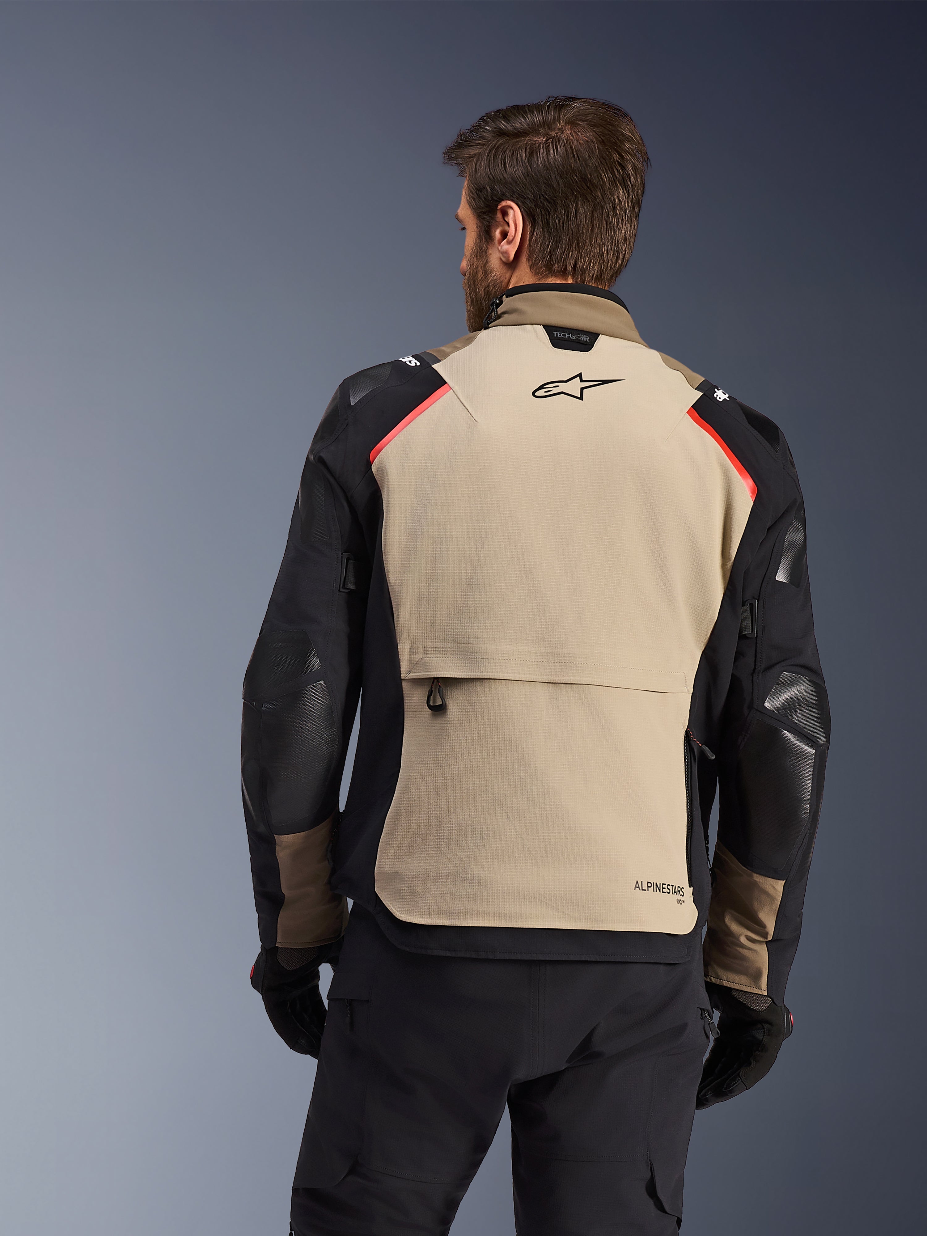 Alpinestars Andes Pro Drystar® XF-jacka, baksida, Walnut och Aluminum, med en stor tanfärgad ryggpanel med svart Alpinestars-logotyp och röda axeldetaljer, stor bakre cargoficka, bärs med svarta byxor och handskar, adventure touring-motorcykelutrustning