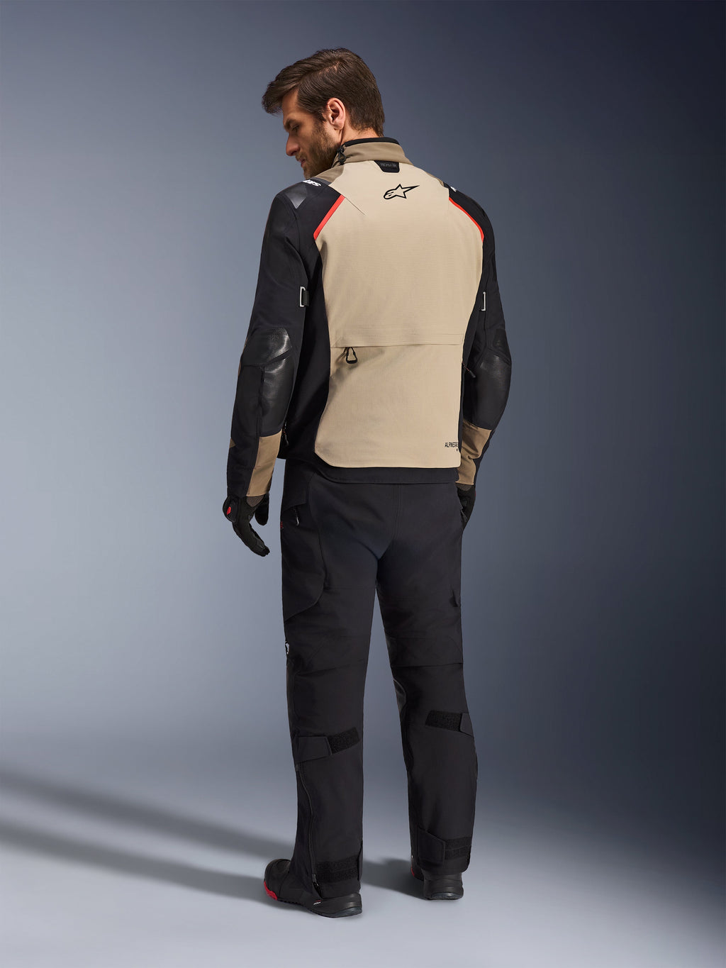 Alpinestars Andes Pro Drystar® XF jacka, motorcykeljacka, valnöt och svart med röda detaljer, sedd bakifrån på modell, adventure touring-design med stor bakre förvaringsficka, bärs med svarta körbyxor och handskar.
