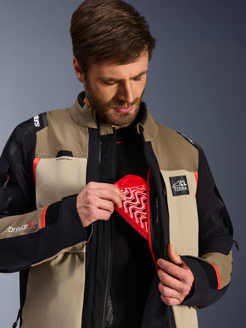 Alpinestars Andes Pro Drystar® XF-jacka i Walnut Aluminum, modell som demonstrerar det röda Nucleon-bröstskyddet i innerfickan, äventyrsmotorcykeljacka med ALL TERRA-logotyp och Drystar XF-logotyper.