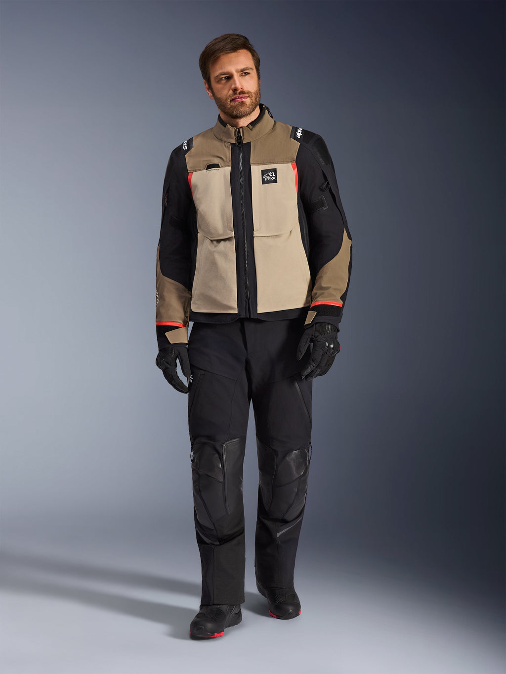 Manlig modell som bär Alpinestars Andes Pro Drystar® XF-jacka i Walnut Aluminum, sandfärgad och svart adventure touring-jacka med röda detaljer, matchad med svarta motorcykelbyxor, handskar och stövlar, helkroppsbild i studio för skyddande körutrustning.