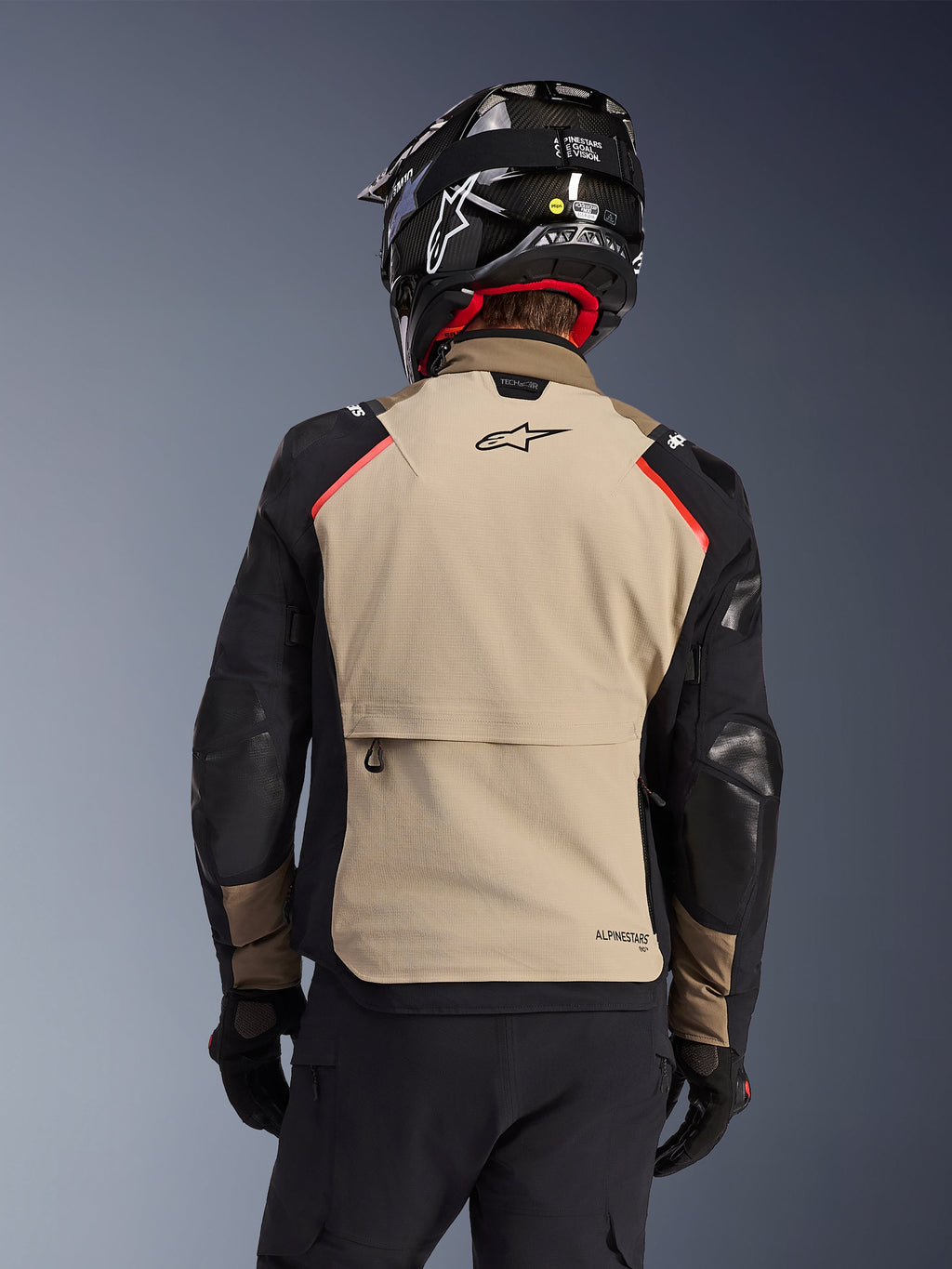 Alpinestars Andes Pro Drystar® XF-jacka i färgen Walnut Aluminum, sedd bakifrån, beige ryggpanel med en stor cargoficka, svarta ärmar med förstärkta paneler, Alpinestars-logotyp på övre delen av ryggen, visas tillsammans med en svart hjälm och handskar för äventyrstouring.