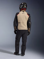 Alpinestars Andes Pro Drystar® XF-jacka i Walnut Aluminum, förare sedd bakifrån, beige ryggpanel med svarta ärmar och röda detaljer, matchad med svarta tekniska adventure-byxor och motorcykelhjälm, förstklassig touringutrustning för alla väder.
