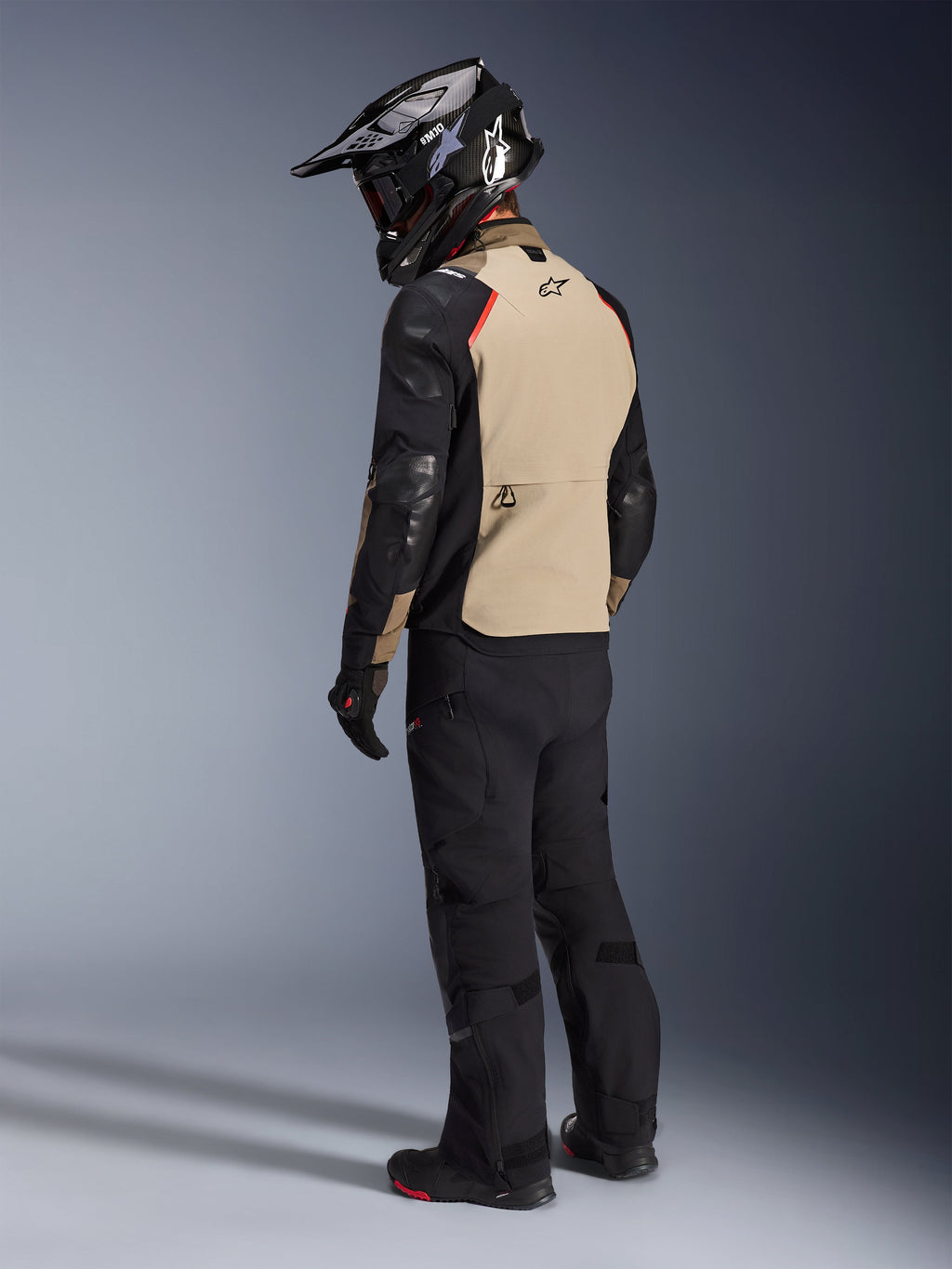 Motorcykelförare sedd bakifrån iförd Alpinestars Andes Pro Drystar® XF Jacket i Walnut Aluminum, med en sandfärgad ryggpanel, svarta ärmar och röda detaljer, svart äventyrshjälm, svarta mc-byxor och svarta stövlar, professionell utrustning för äventyrstouring.