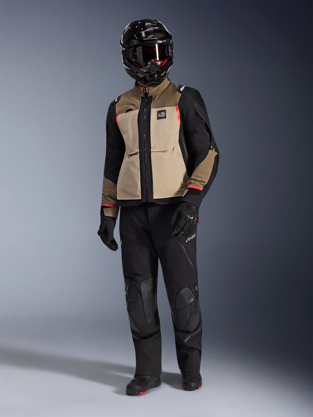Alpinestars Andes Pro Drystar® XF-jacka i Walnut Aluminum, äventyrsmotorcykeljacka buren med matchande svarta byxor, en svart äventyrshjälm med goggles och svarta stövlar, med beigea och svarta paneler och röda detaljer.