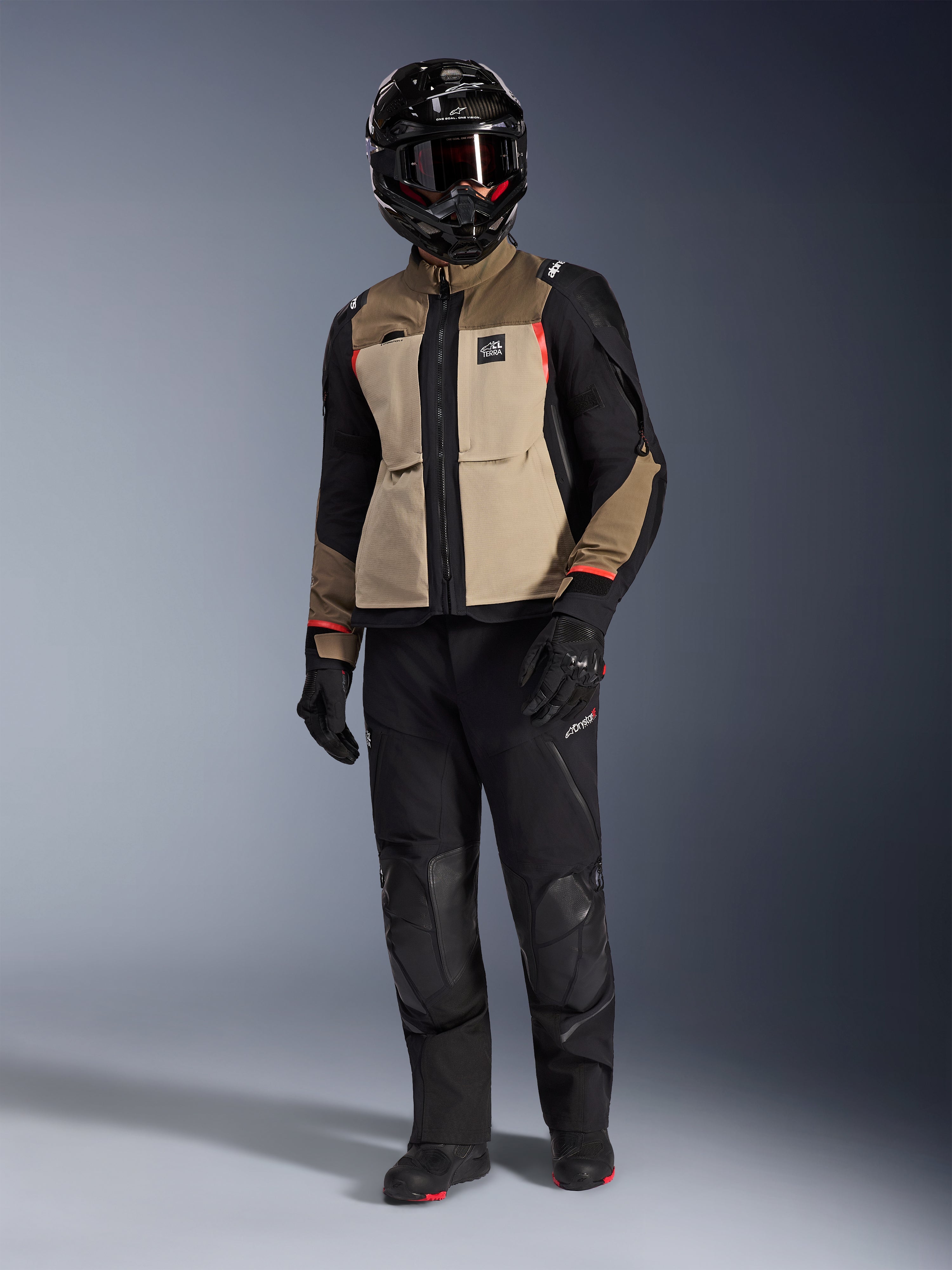 Alpinestars Andes Pro Drystar® XF-jacka i Walnut Aluminum, äventyrsmotorcykeljacka buren med matchande svarta byxor, en svart äventyrshjälm med goggles och svarta stövlar, med beigea och svarta paneler och röda detaljer.