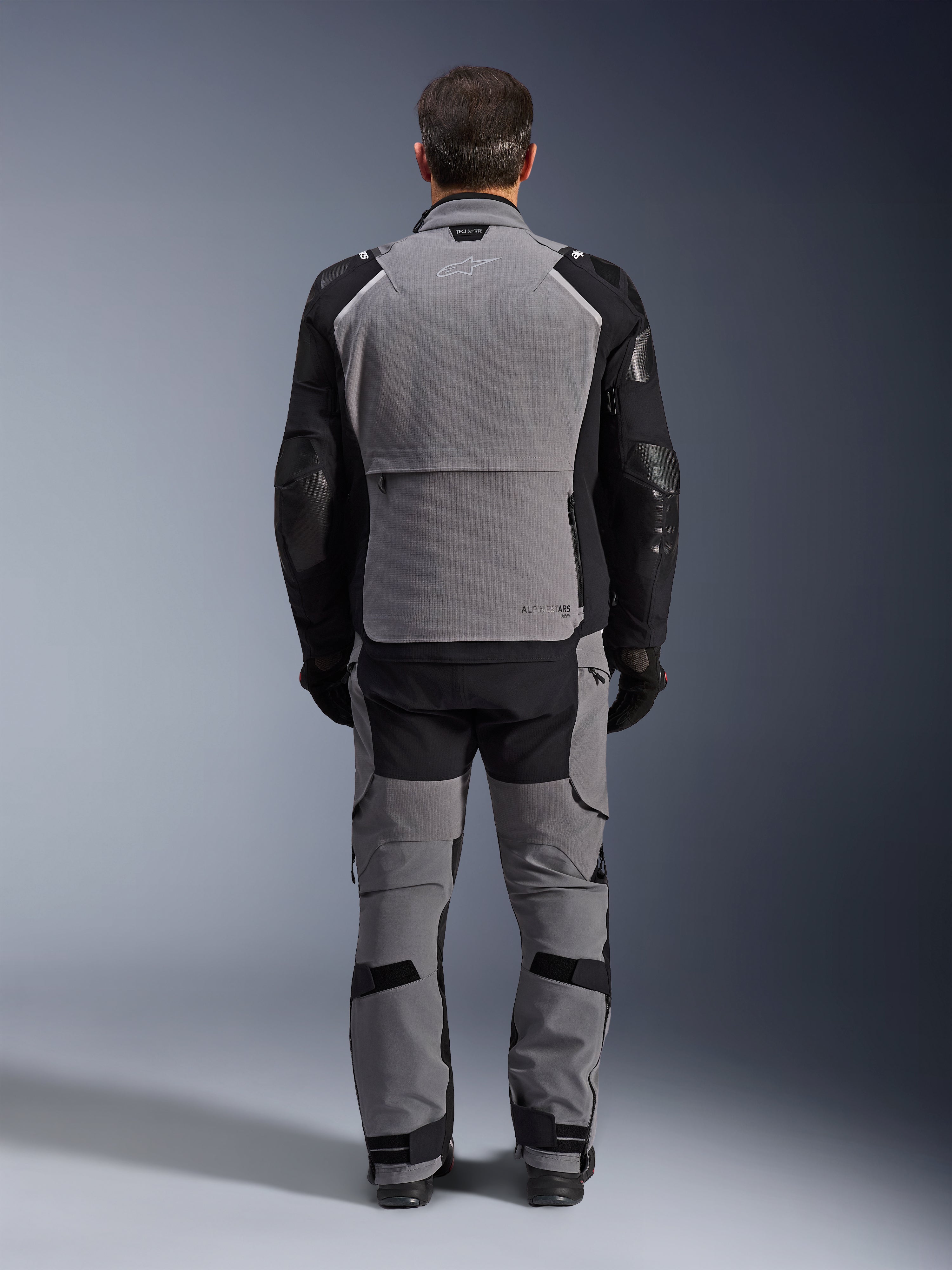 Alpinestars Andes Pro Drystar® XF Jacka, Dark Gray Black, baksida av en förare som bär matchande byxor, äventyrstouring-motorcykeljacka med en stor grå ryggpanel, svarta förböjda ärmar med skydd och ett bakre förvaringsfack