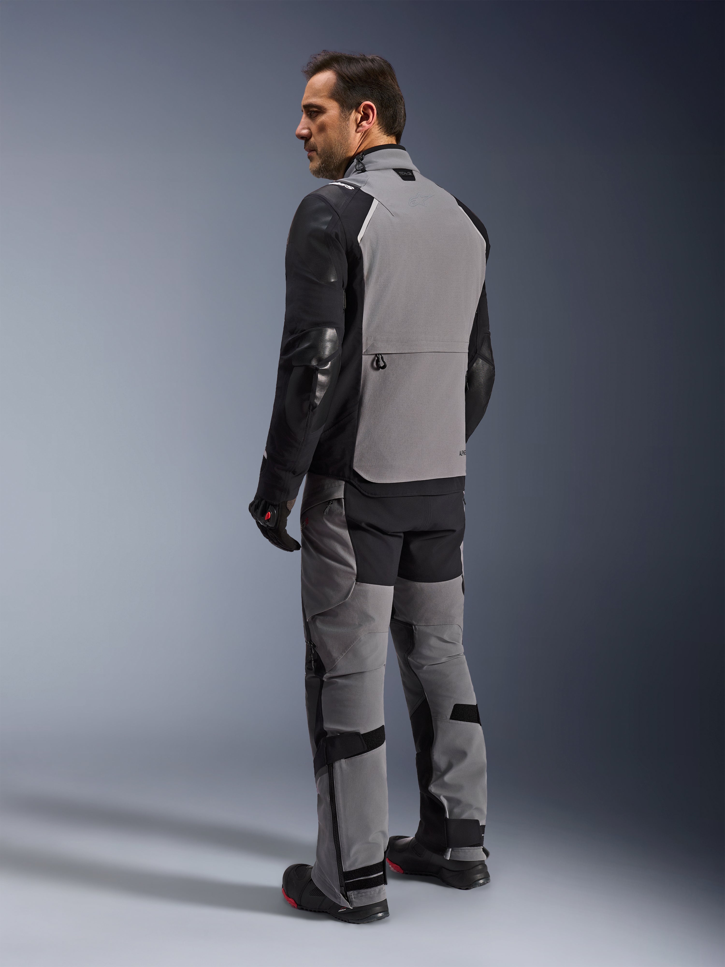 Alpinestars Andes Pro Drystar® XF-jacka, äventyrstouring-motorcykeljacka i mörkgrått och svart, vy bakifrån på en modell som visar den stora ryggpanelen och förstärkta ärmar, kombinerad med matchande äventyrsbyxor, handskar och stövlar.