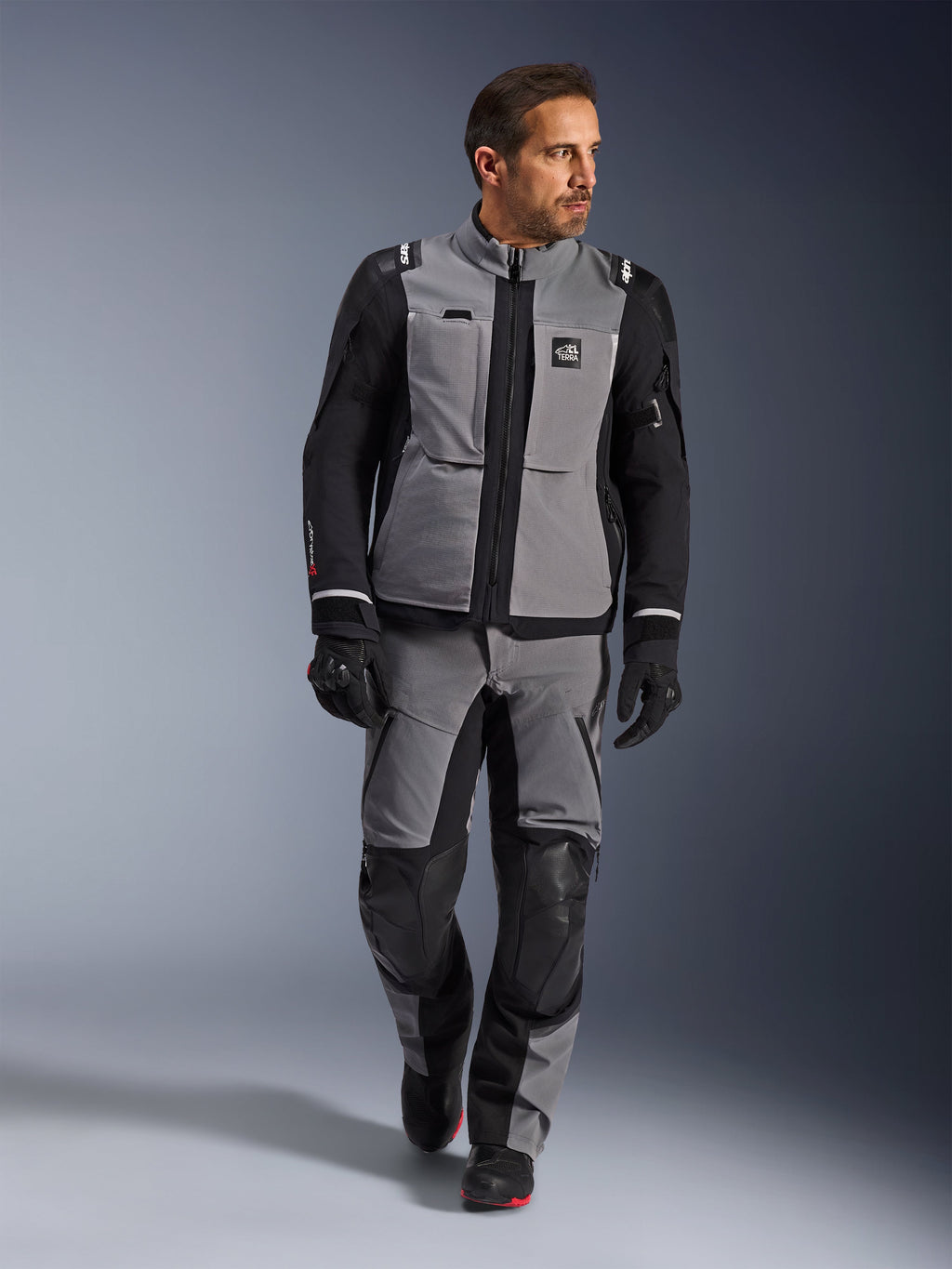 Manlig modell iförd Alpinestars Andes Pro Drystar® XF-jacka i mörkgrått och svart, motorcykelställ för äventyrskörning med matchande byxor och handskar, tekniskt textilskal med 'All Terra'-märkning, komplett utrustning för långdistansäventyrstouring