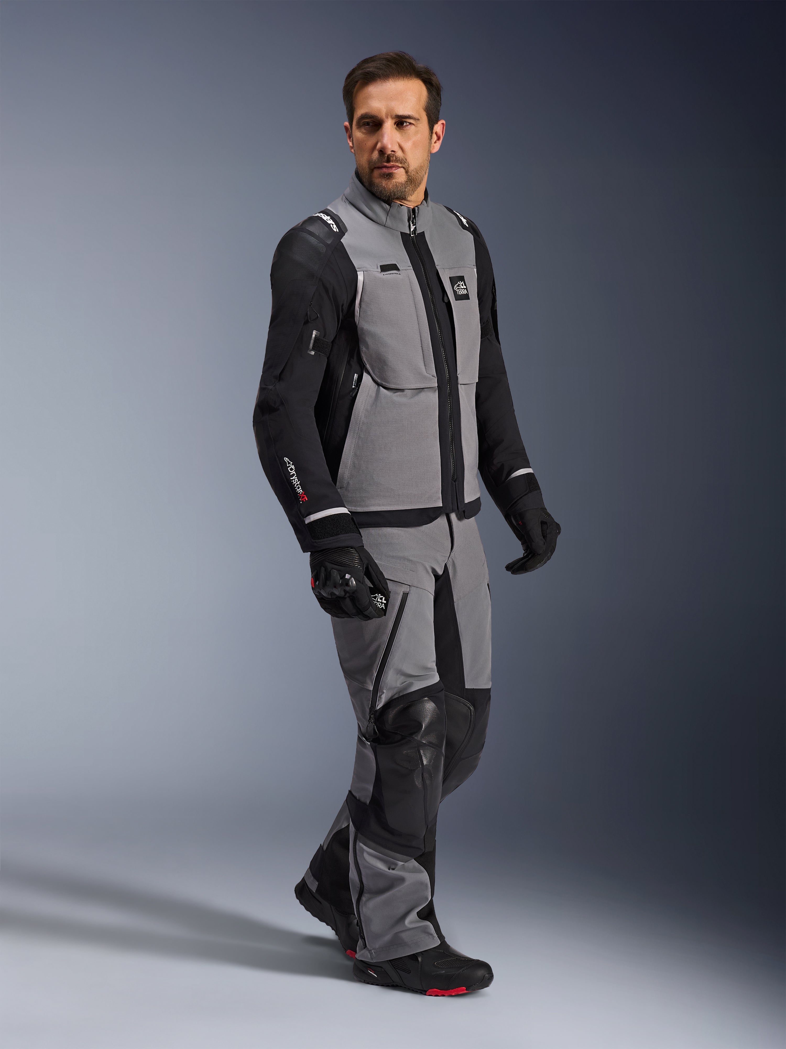 Alpinestars Andes Pro Drystar® XF-jacka, äventyrsmotorcykeljacka, mörkgrå och svart, bärs av en modell med matchande textilbyxor och svarta handskar, design med flera fickor, tekniskt 3-lagers laminerat skal och artikulerad passform för långdistansäventyrskörning.