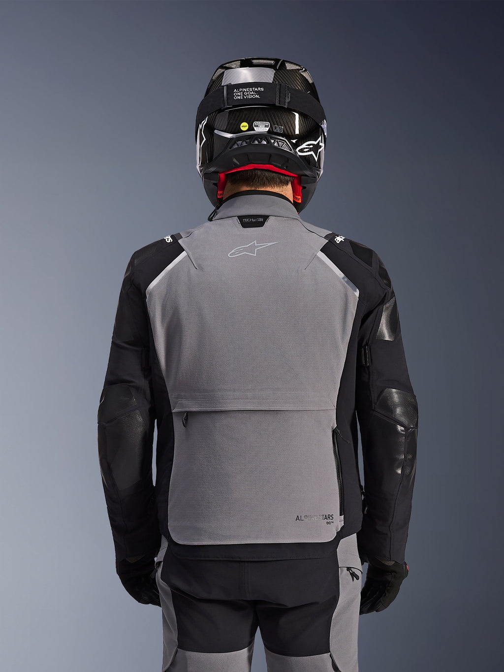 Alpinestars Andes Pro Drystar® XF-jacka, mörkgrå och svart, baksida som visar en stor bakre förvaringsficka och Tech-Air-märkning, bärs av en förare med svart hjälm och matchande mc-byxor, professionell äventyrstouringutrustning