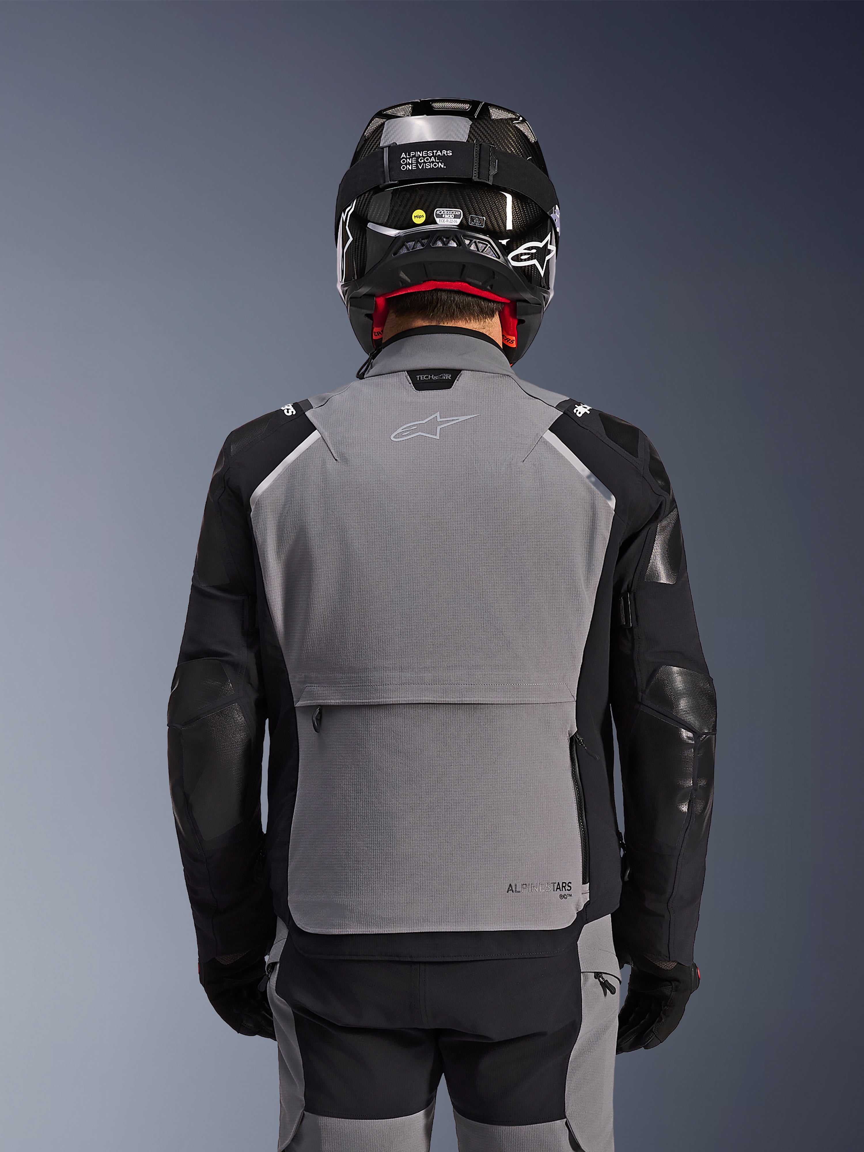 Alpinestars Andes Pro Drystar® XF-jacka, mörkgrå och svart, baksida som visar en stor bakre förvaringsficka och Tech-Air-märkning, bärs av en förare med svart hjälm och matchande mc-byxor, professionell äventyrstouringutrustning