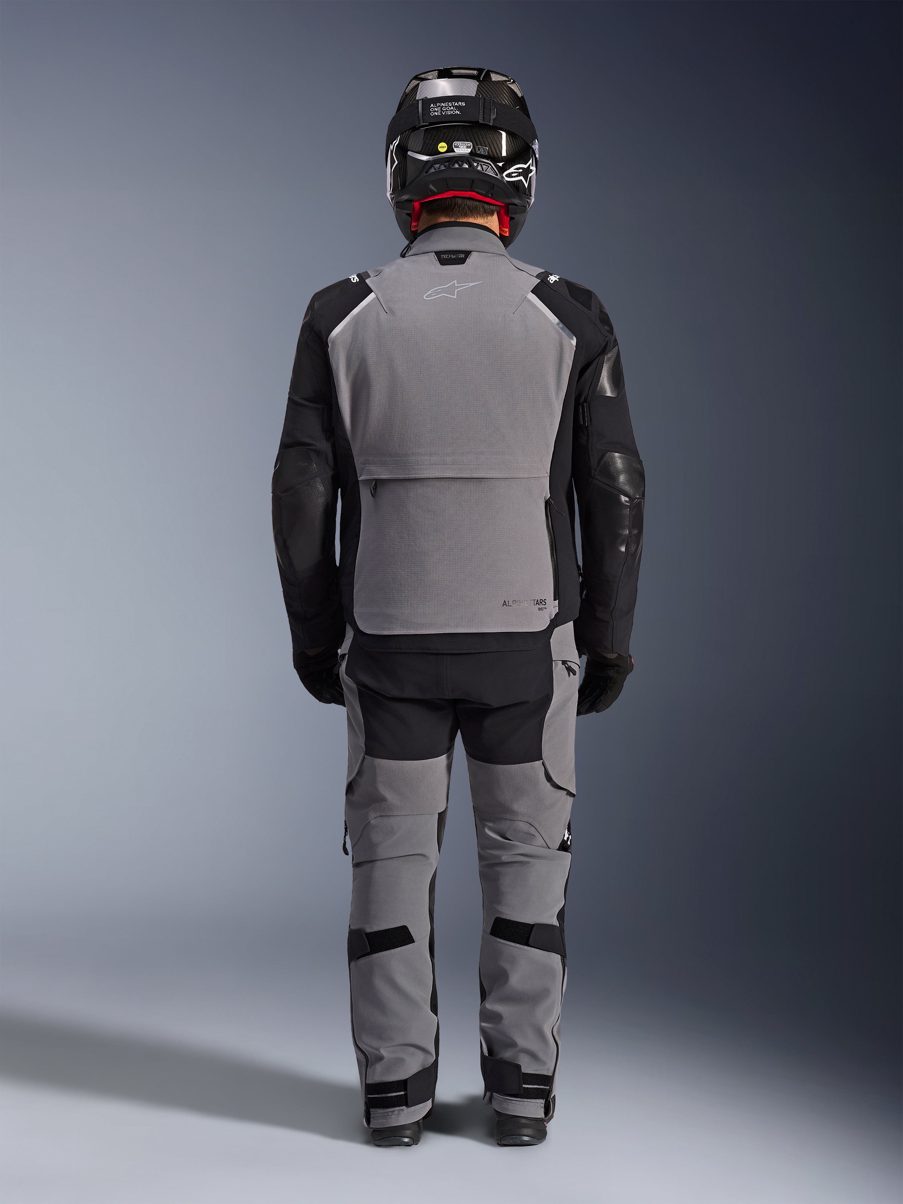 Alpinestars Andes Pro Drystar® XF-jacka, baksida, mörkgrå och svart adventure touring-utrustning, bärs med matchande grå och svarta byxor, svart kolfiberhjälm och svarta handskar, med tekniska paneler och reflekterande logotyper